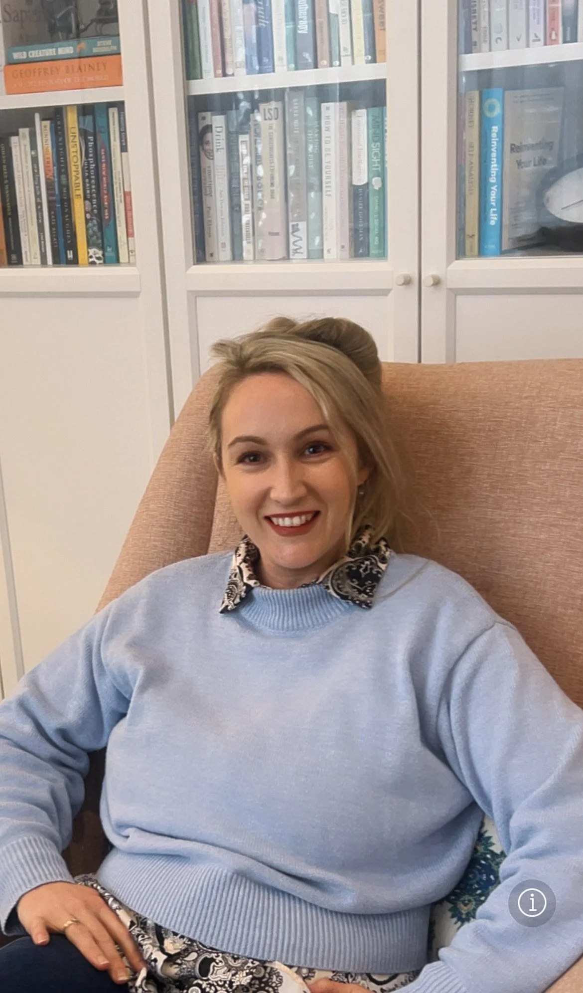 A woman with blonde hair smiling, sitting in a pink upholstered armchair, wearing a light blue sweater over a beige and black patterned shirt, with a bookshelf filled with books behind her.