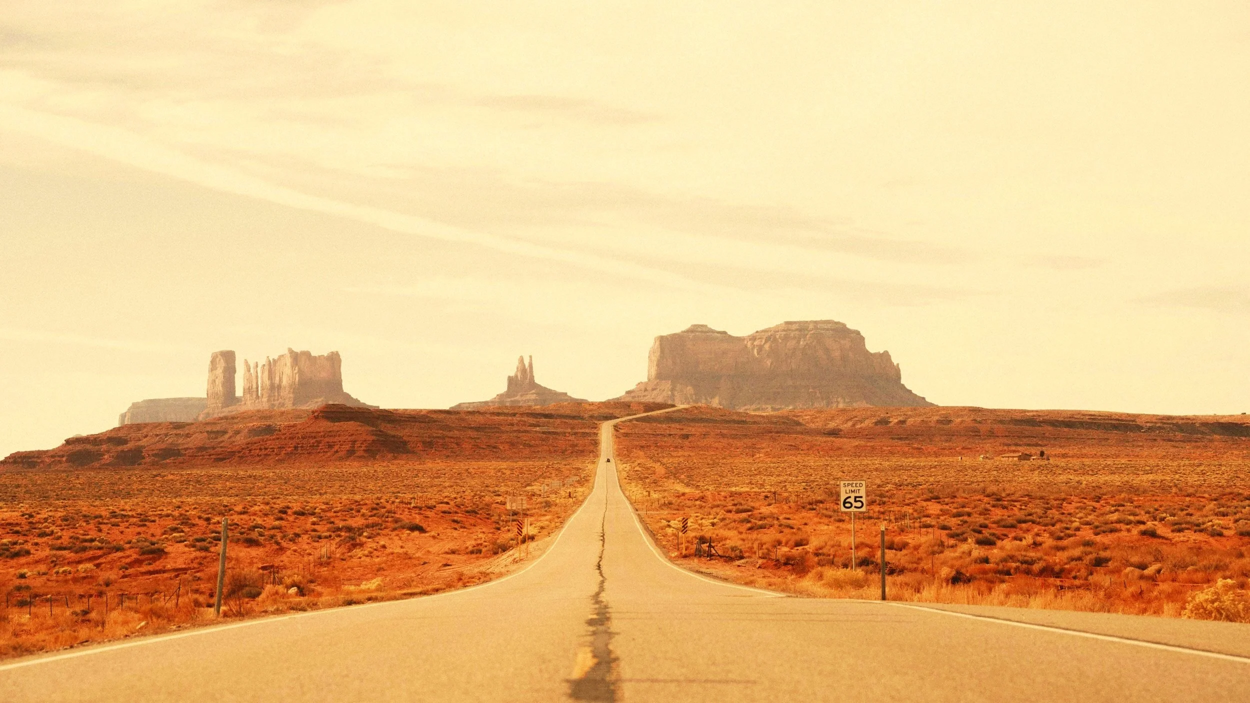 A straight road leading through a desert landscape with rock formations in the background and a speed limit sign of 65 miles per hour.