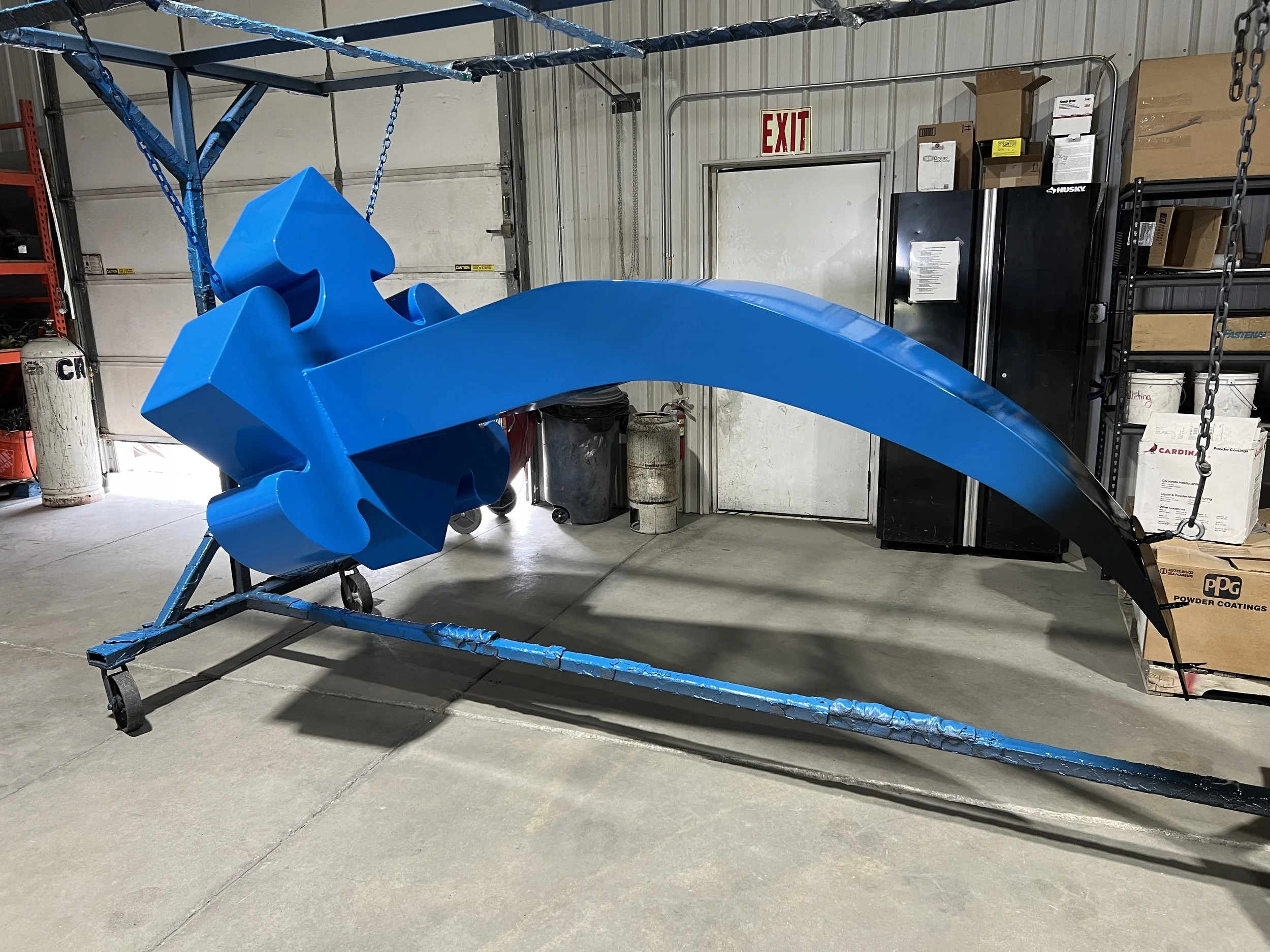 Large blue and black sculpture or airplane nose cone with a sword shape on top, mounted on a blue metal stand in a warehouse or workshop.