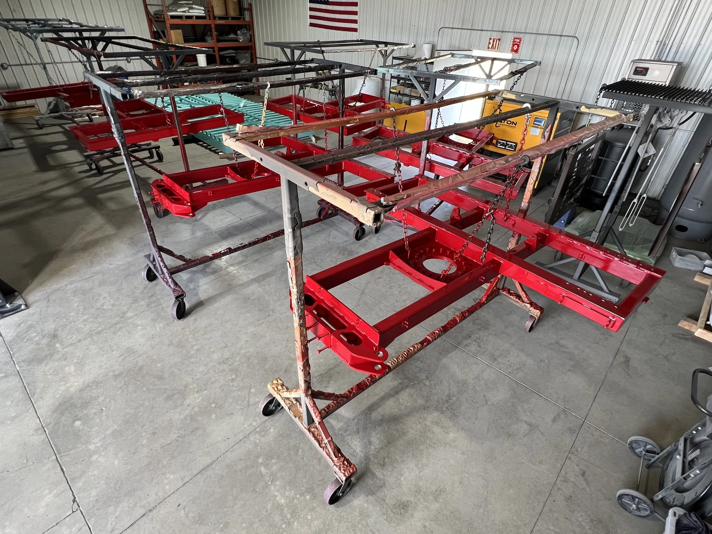 Industrial workshop with multiple red metal frames on wheeled carts, some with chains and top bars, inside a storage facility with metal walls and shelves.