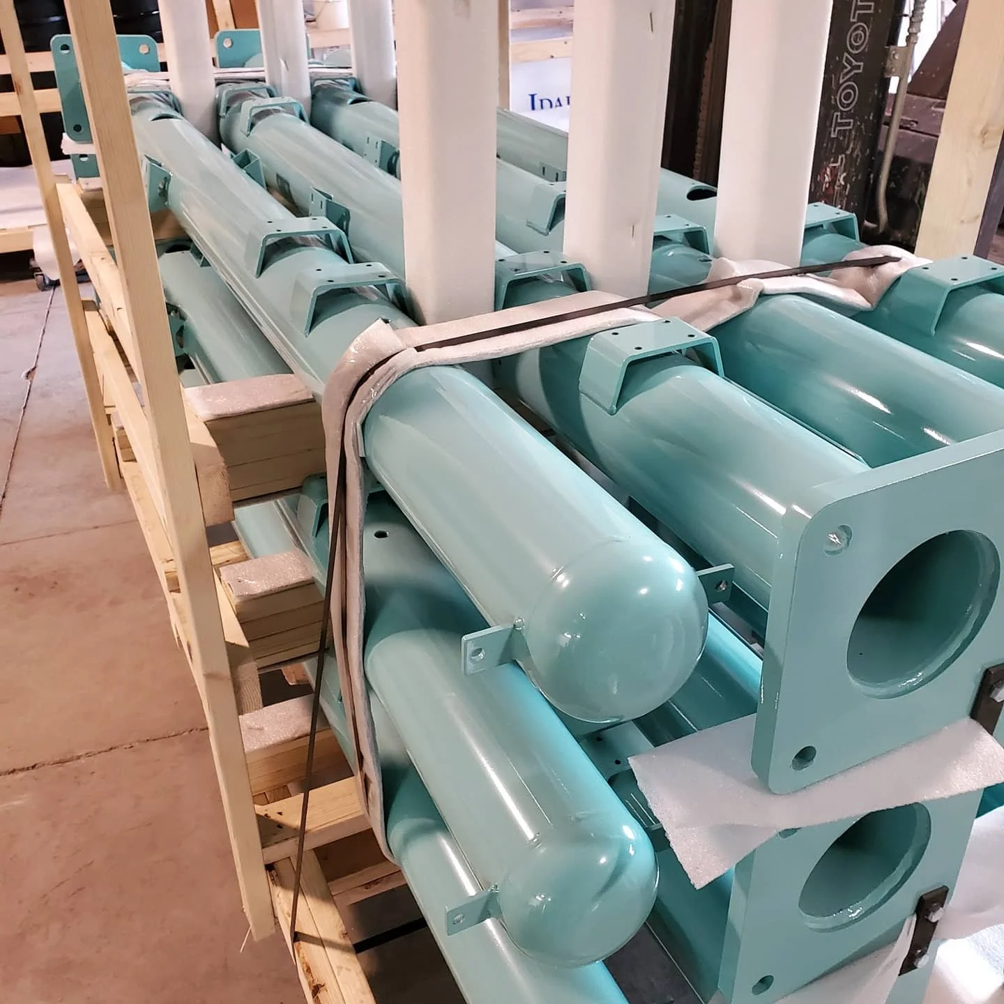 Multiple teal-colored metal pipes and components stacked on a wooden pallet, likely parts for industrial equipment or machinery.