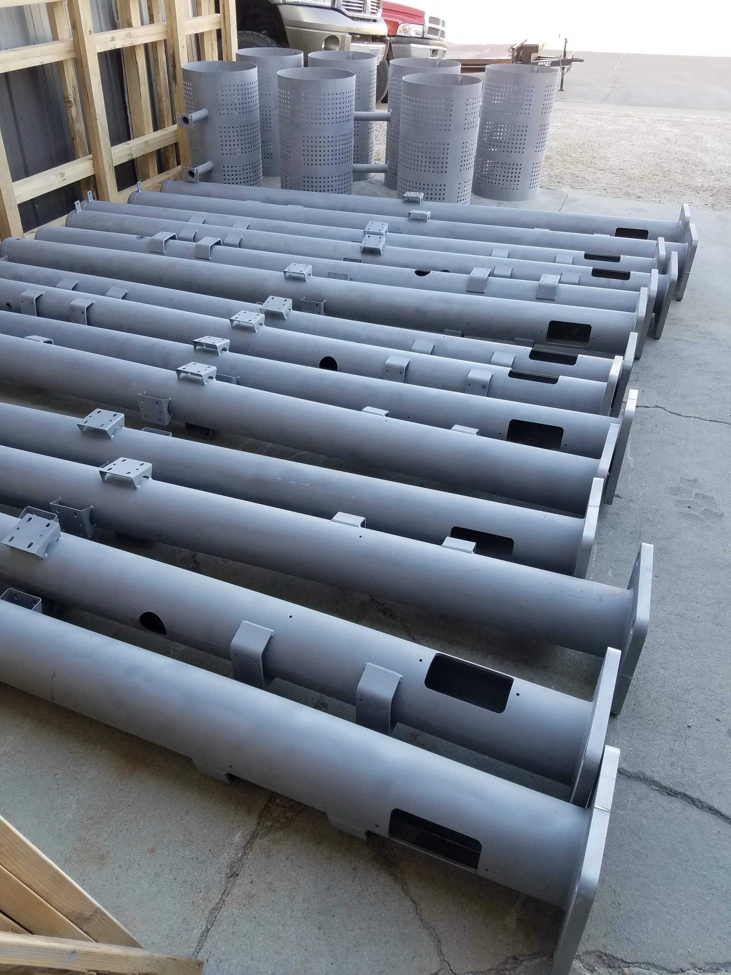 Row of large gray metal poles with brackets and rectangular openings, and cylindrical metal structures with perforated patterns in the background, outdoors on a concrete surface.