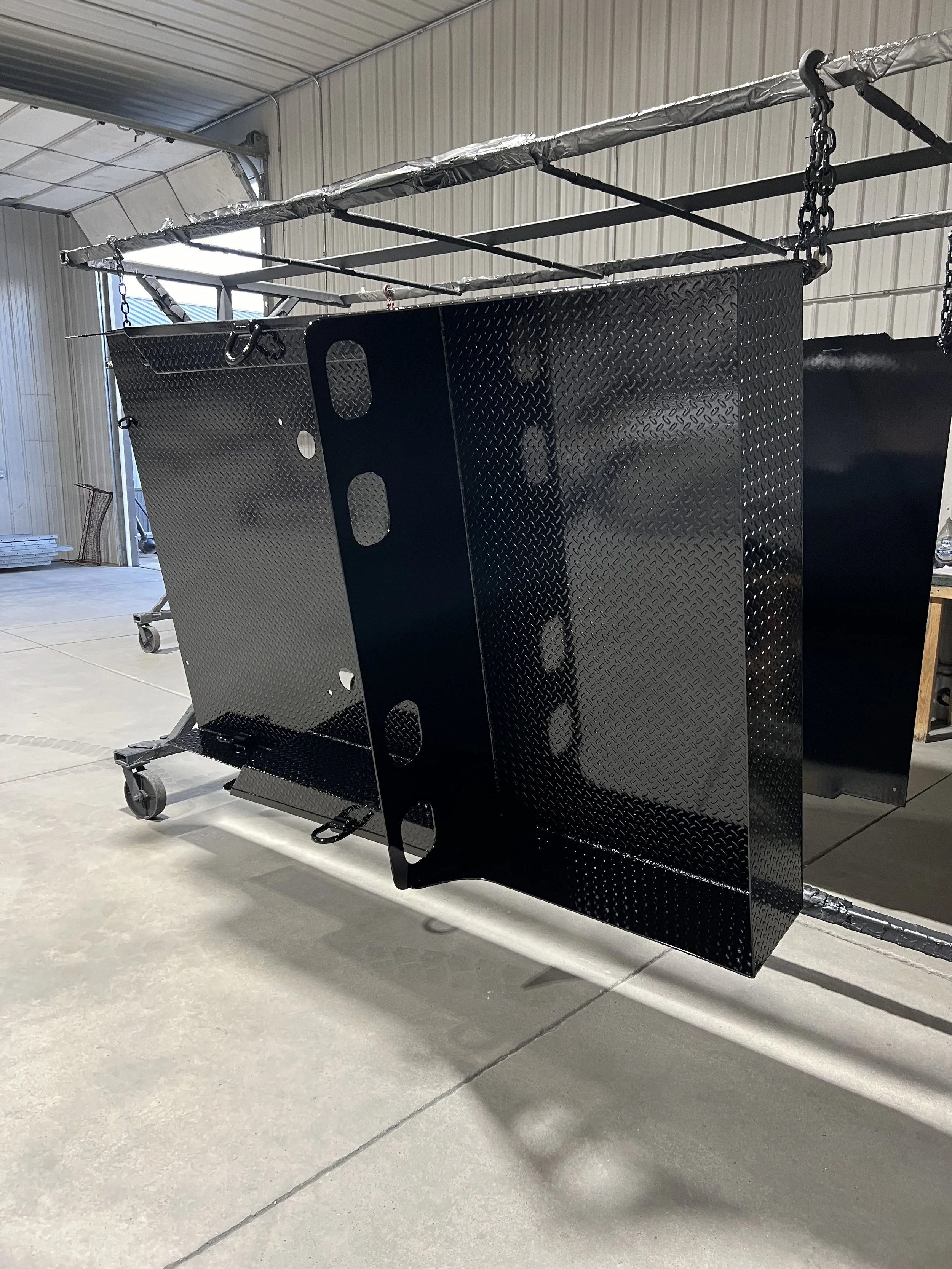 A black metal skid plate with a diamond plate pattern is hanging from an overhead support in a warehouse or workshop setting.