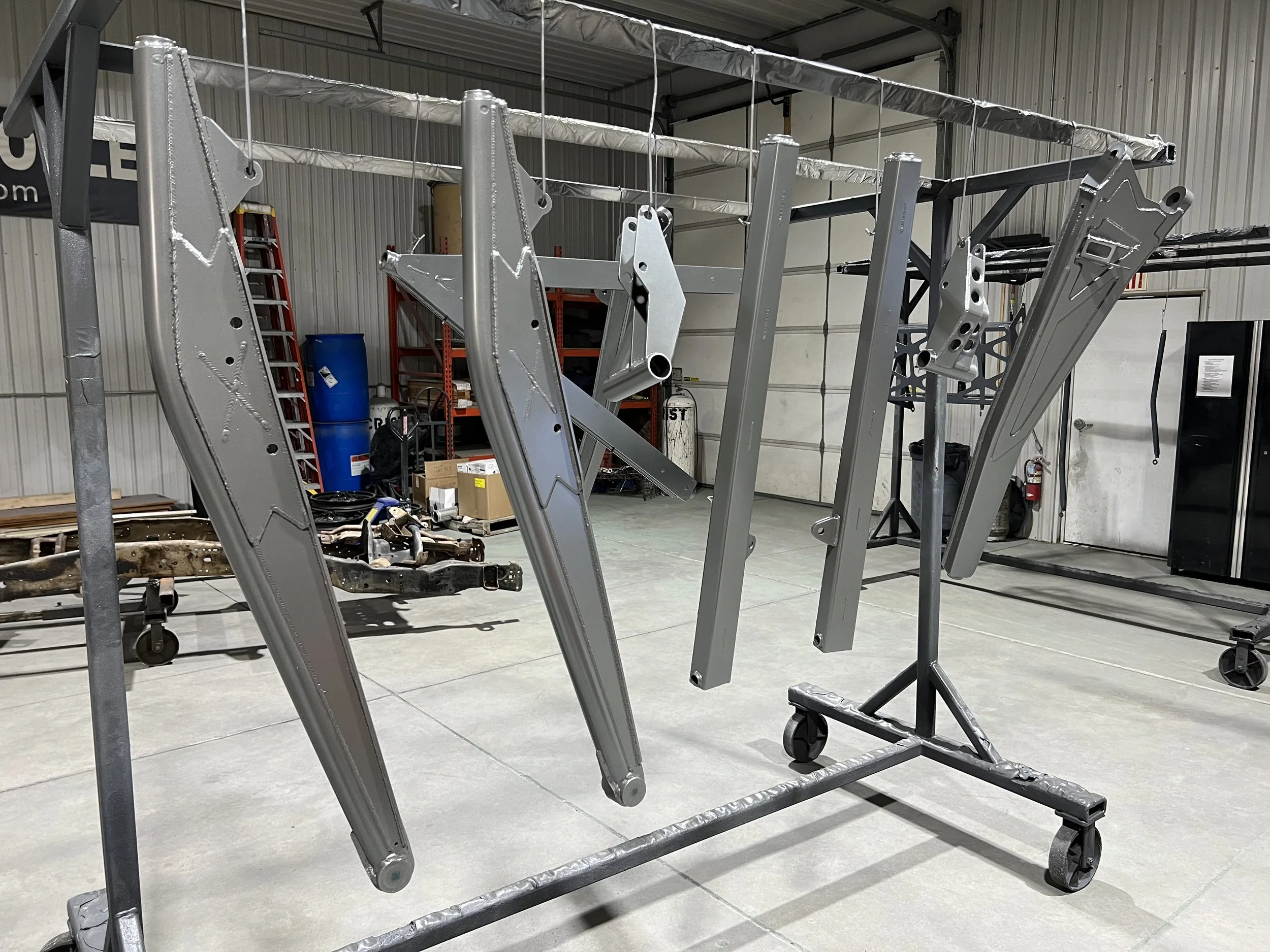Metal parts hanging on a rack in a warehouse or workshop, with a background featuring storage shelves, tools, and a fire extinguisher.