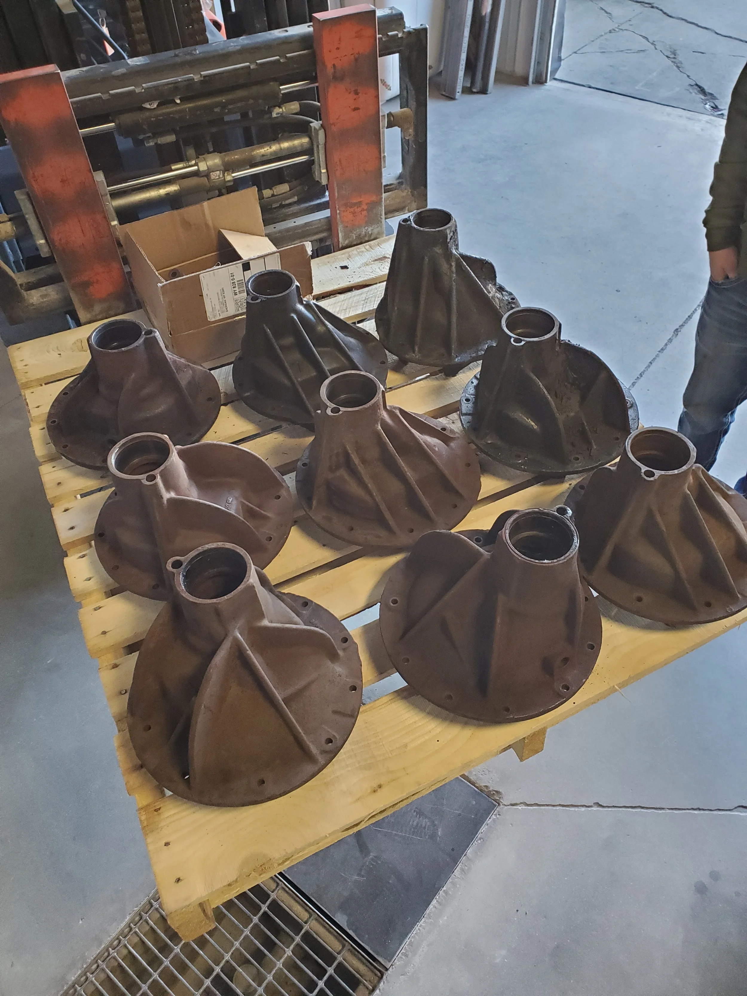 Eight large, rusty metal hub housings are arranged on a wooden workbench in a workshop. A cardboard box and industrial machinery are visible in the background, along with part of a person's arm and leg on the right side.