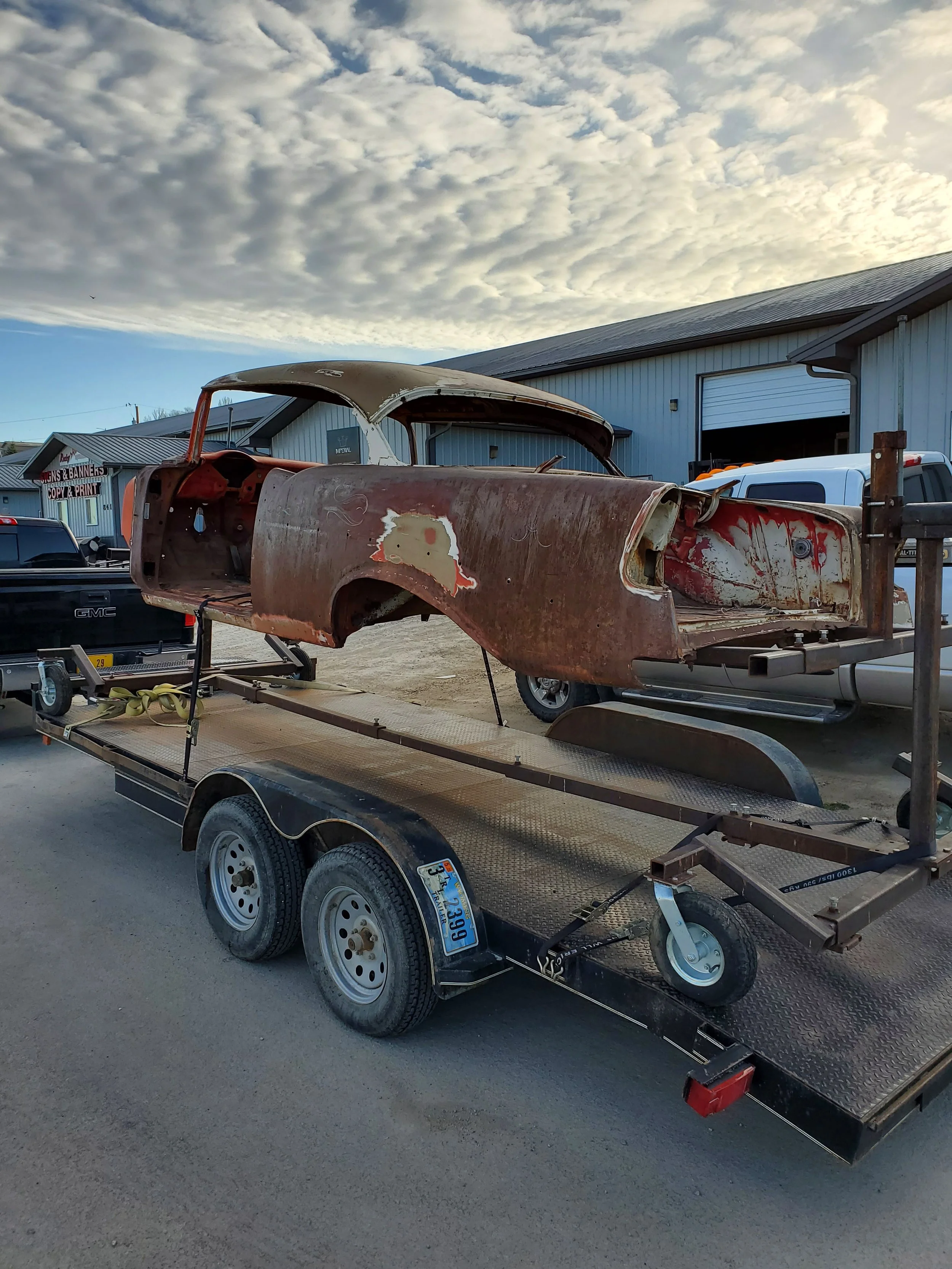 Old rusty car body on a flatbed trailer outside a building with cloudy sky overhead.