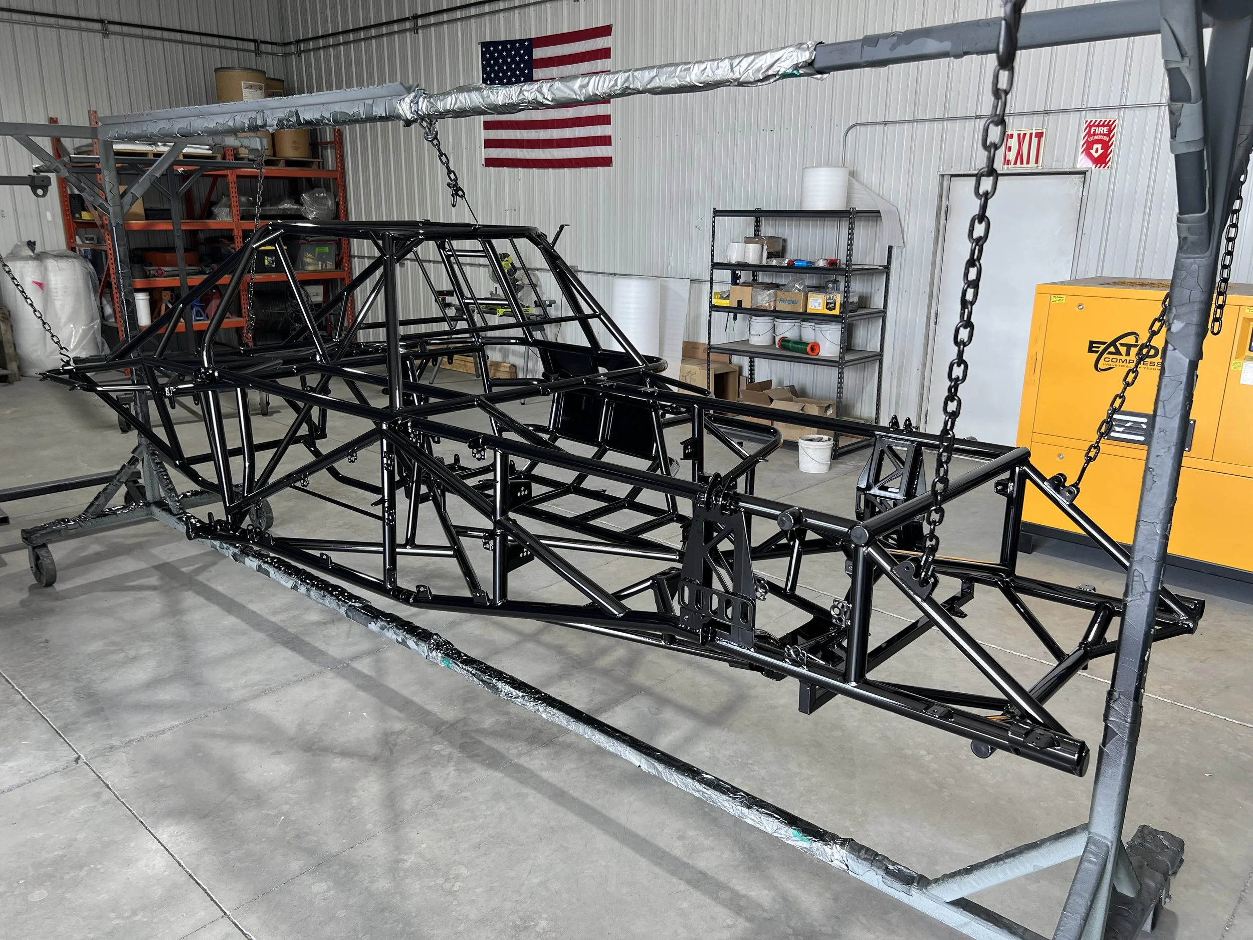 A black metal race car chassis on a metal stand inside a workshop with tools and equipment, including a yellow compressor, a shelf with parts, and an American flag.