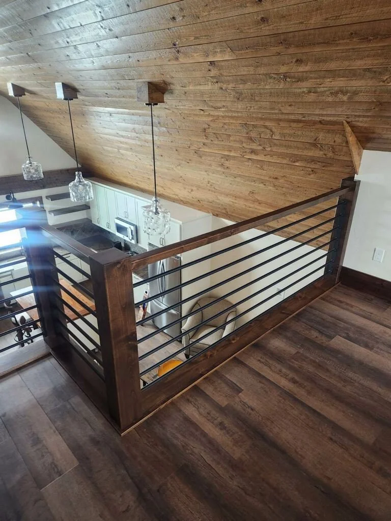 View of a loft area overlooking a kitchen with white cabinets, microwave, and hanging pendant lights, featuring a wooden railing and flooring, and a sloped wooden ceiling.