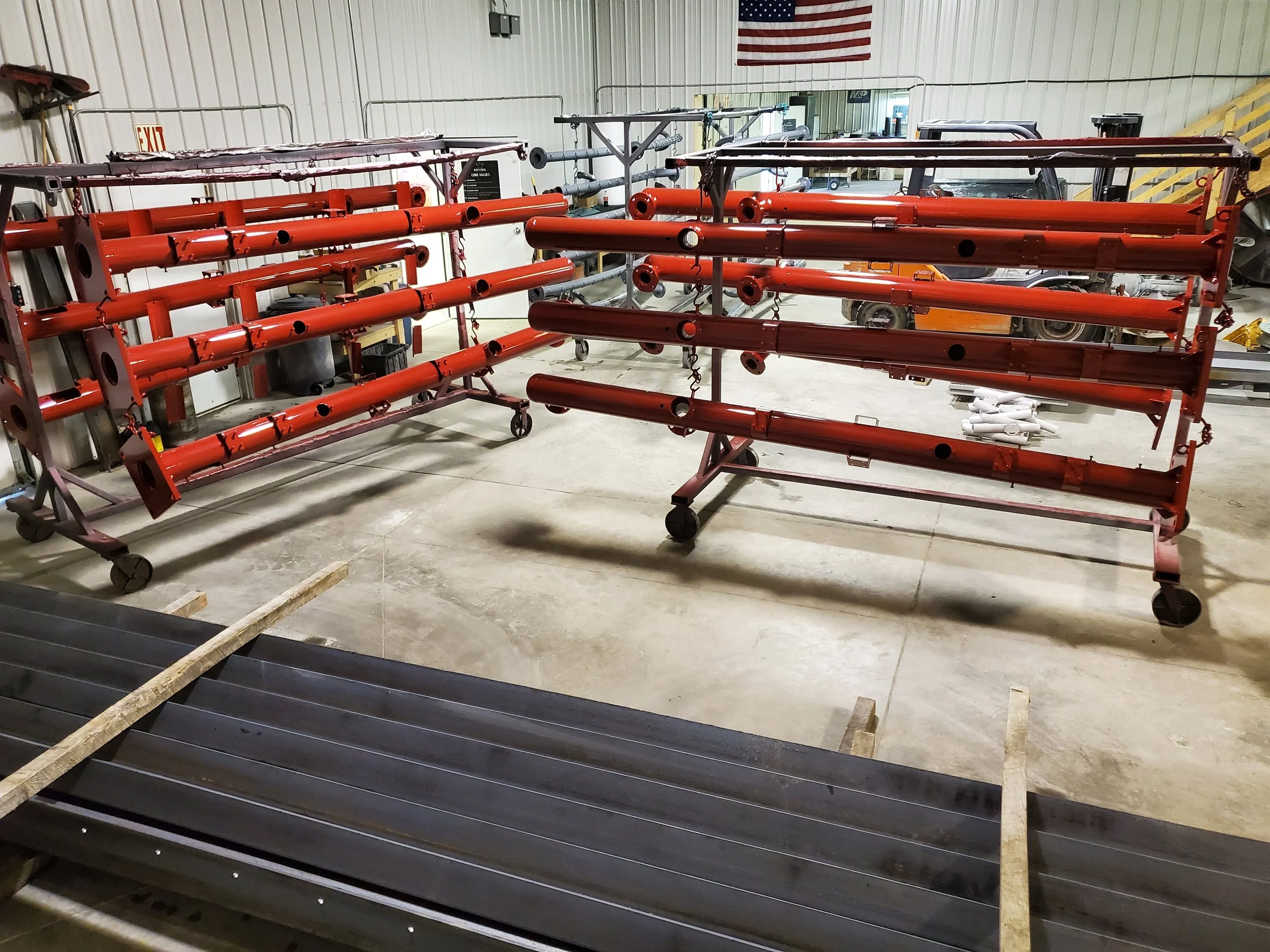 Two red metal frames with horizontal bars and wheels in a warehouse or workshop setting, with some white objects on the floor and vehicles in the background.