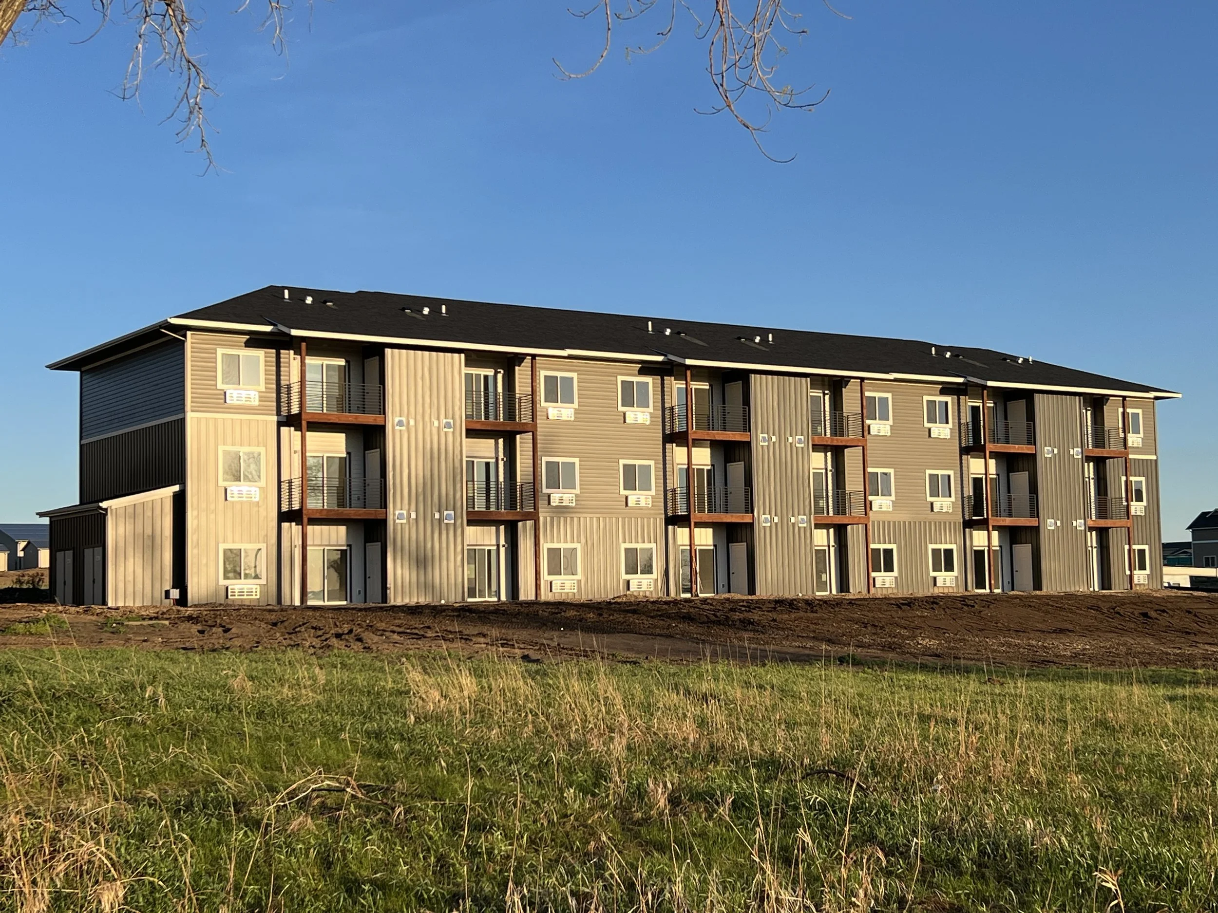 A modern, multi-story residential building with multiple balconies and numerous windows, situated on a grassy area with a clear blue sky in the background.