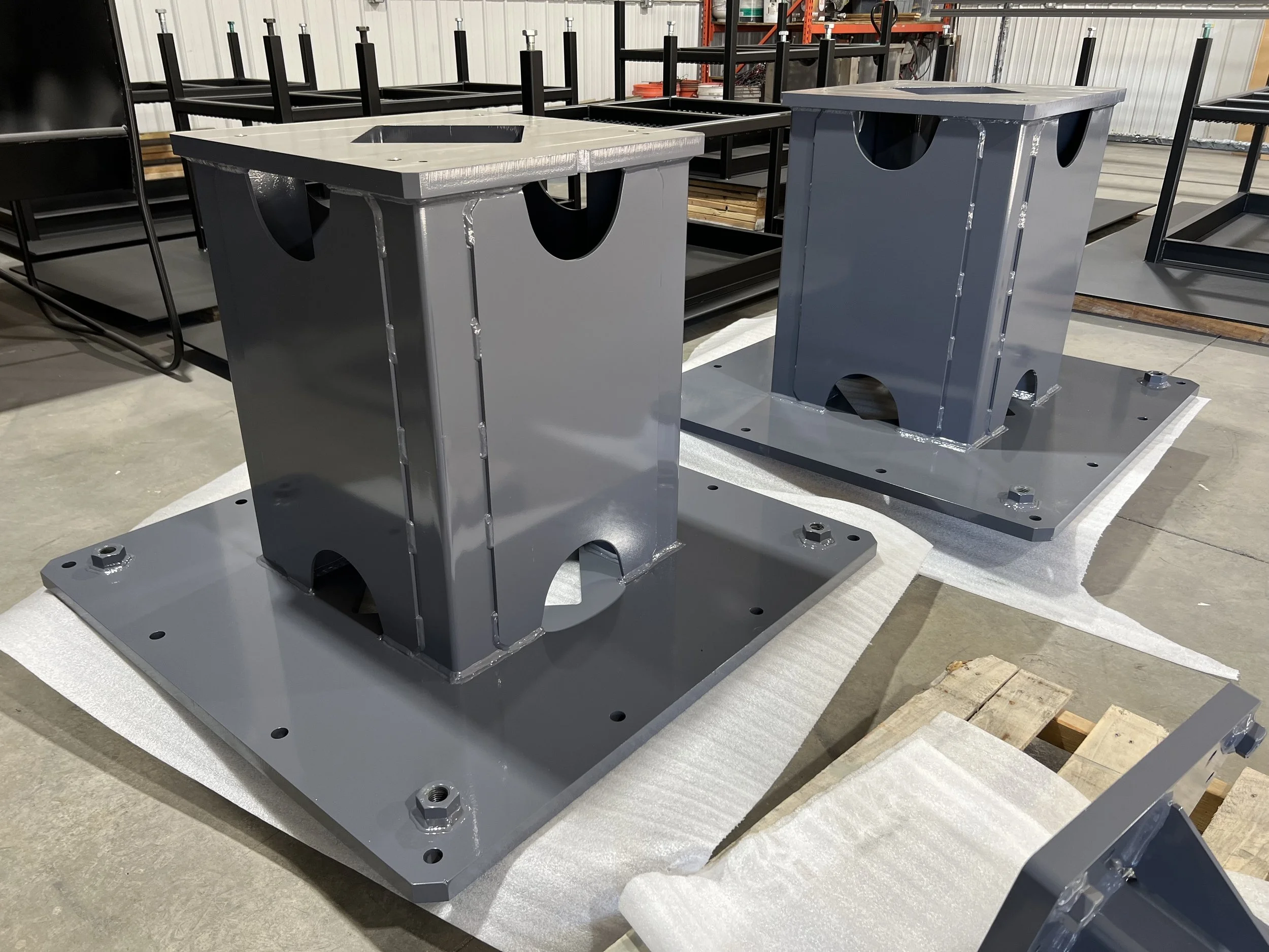 Two large gray metal bases with vertical supports and cutouts, placed on white foam padding in a warehouse or factory setting.