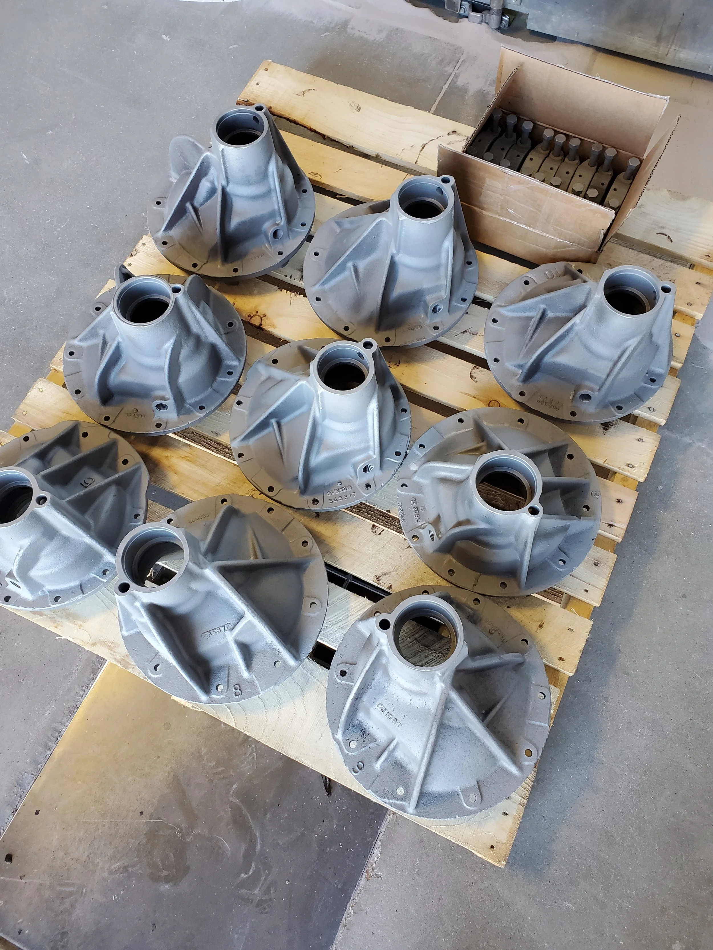 Metal automotive parts, likely differential covers, arranged on wooden pallets with a cardboard box containing small metal components nearby.
