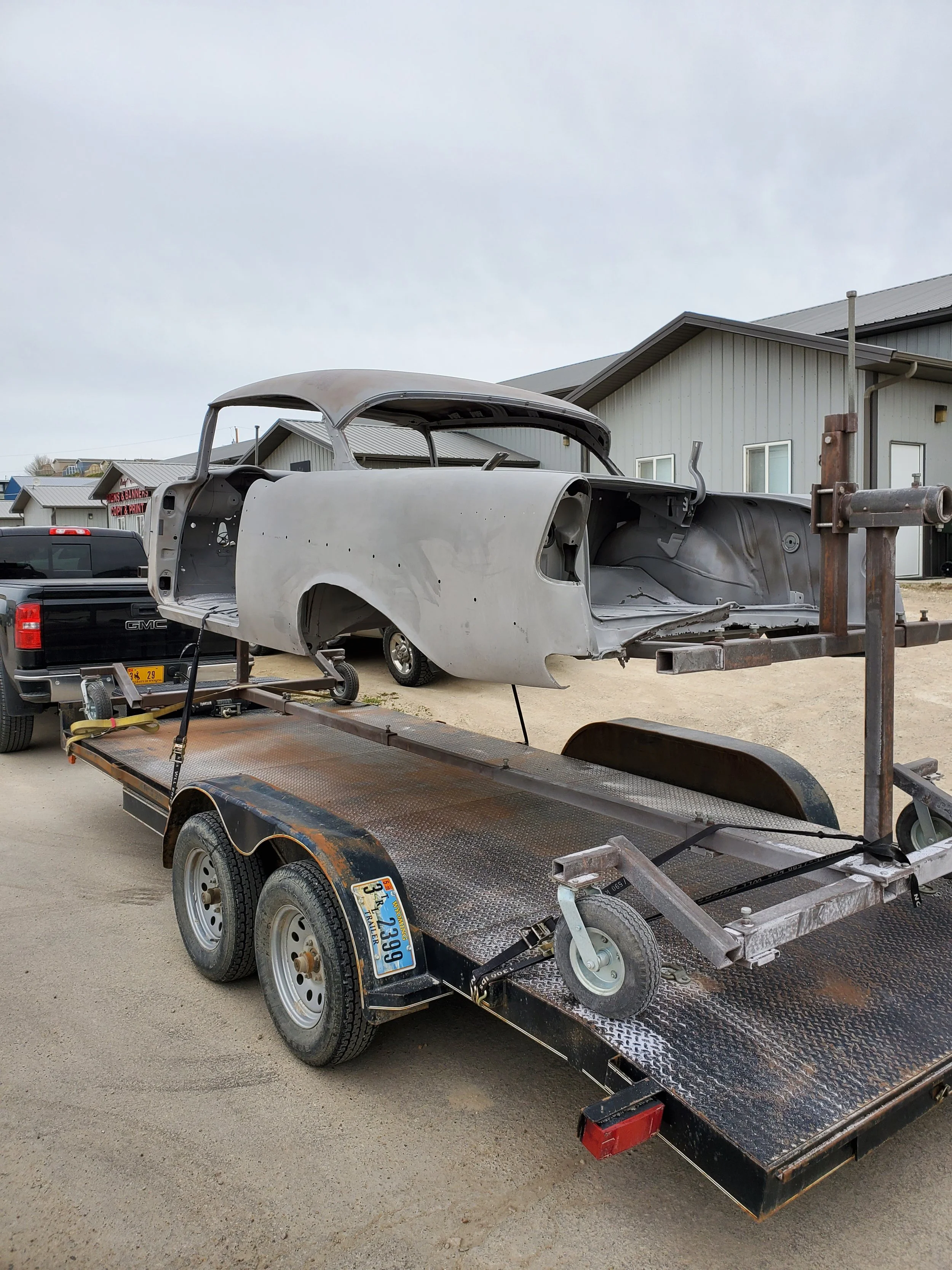 A classic car body shell mounted on a flatbed trailer, ready for transport. The car shell is gray and in incomplete condition, with no windows or wheels. The trailer is parked outdoors near residential or commercial buildings.