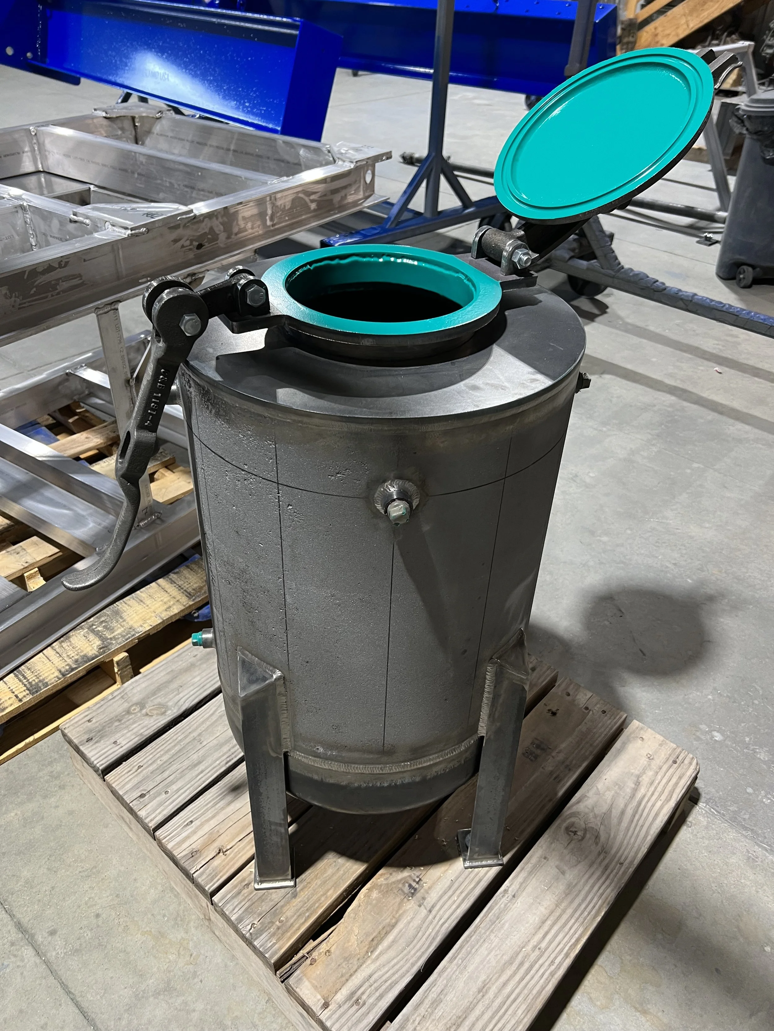 A stainless steel, cylindrical industrial trash can with a teal lid and opening, placed on a wooden pallet in a factory or warehouse setting.