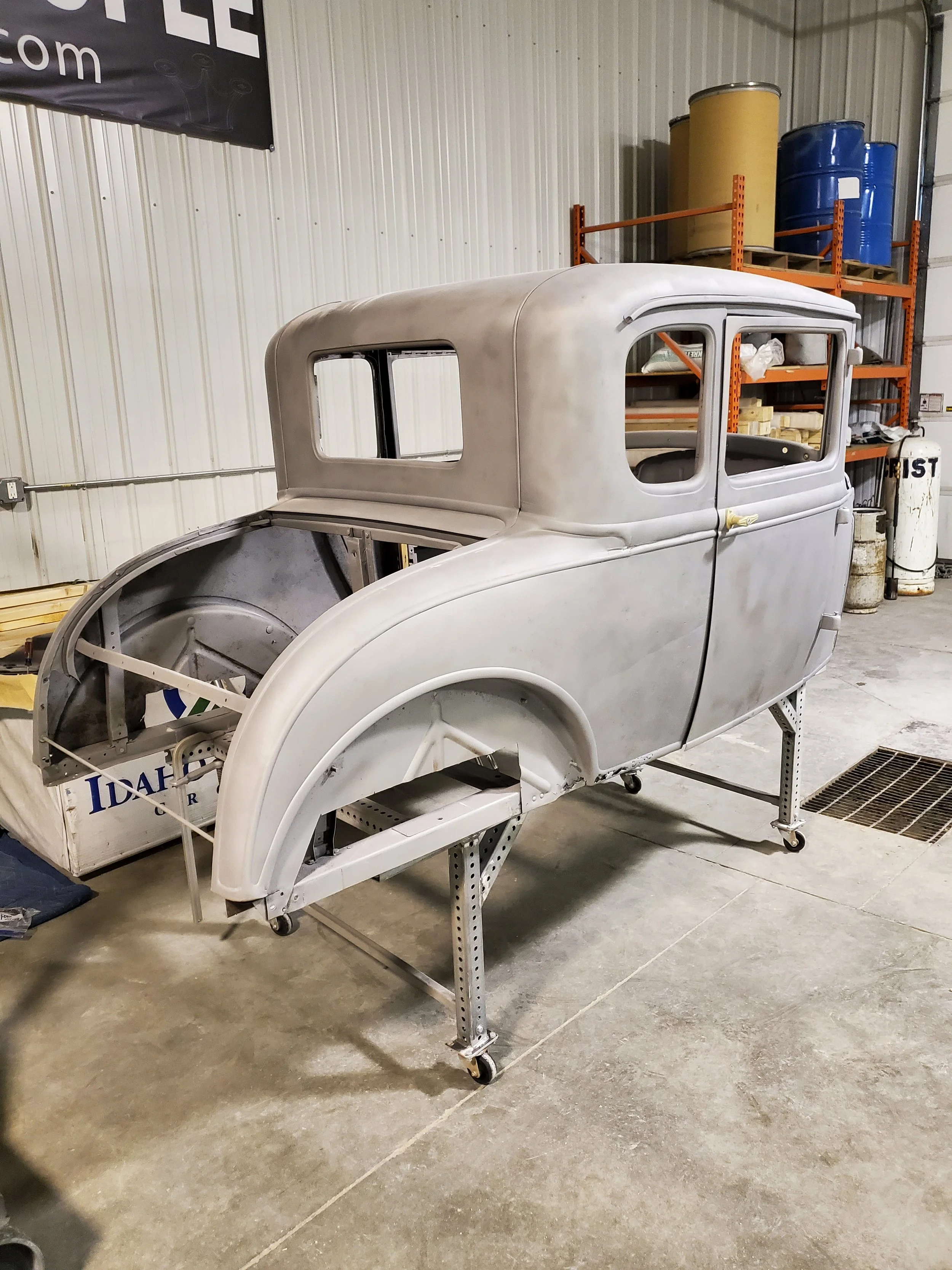 A vintage car body on a metal stand inside a workshop, with shelves holding barrels and supplies in the background.