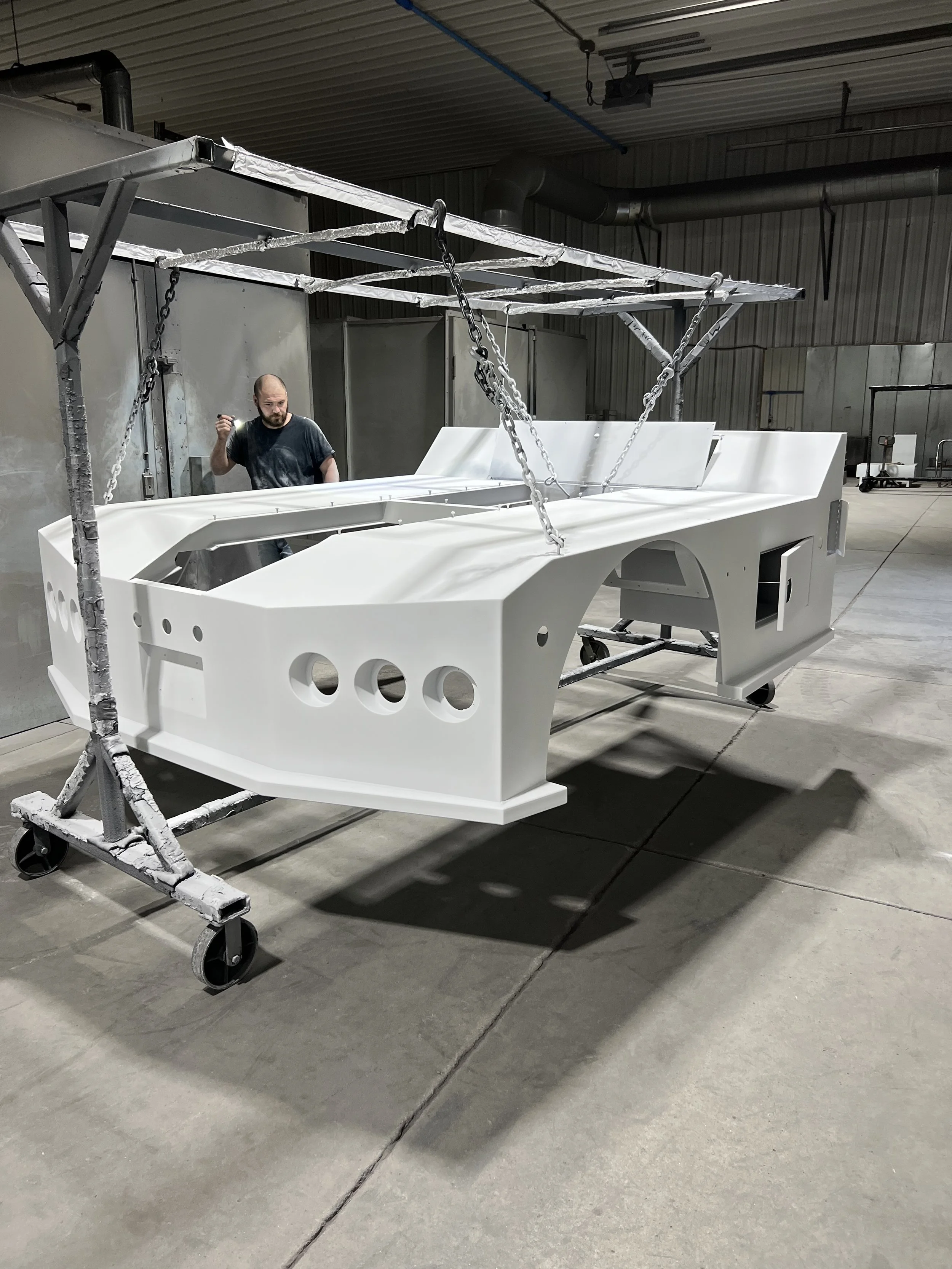A large white fiberglass boat hull is mounted on a wheeled metal frame inside a manufacturing or assembly warehouse. A worker stands nearby inspecting or working on the hull.