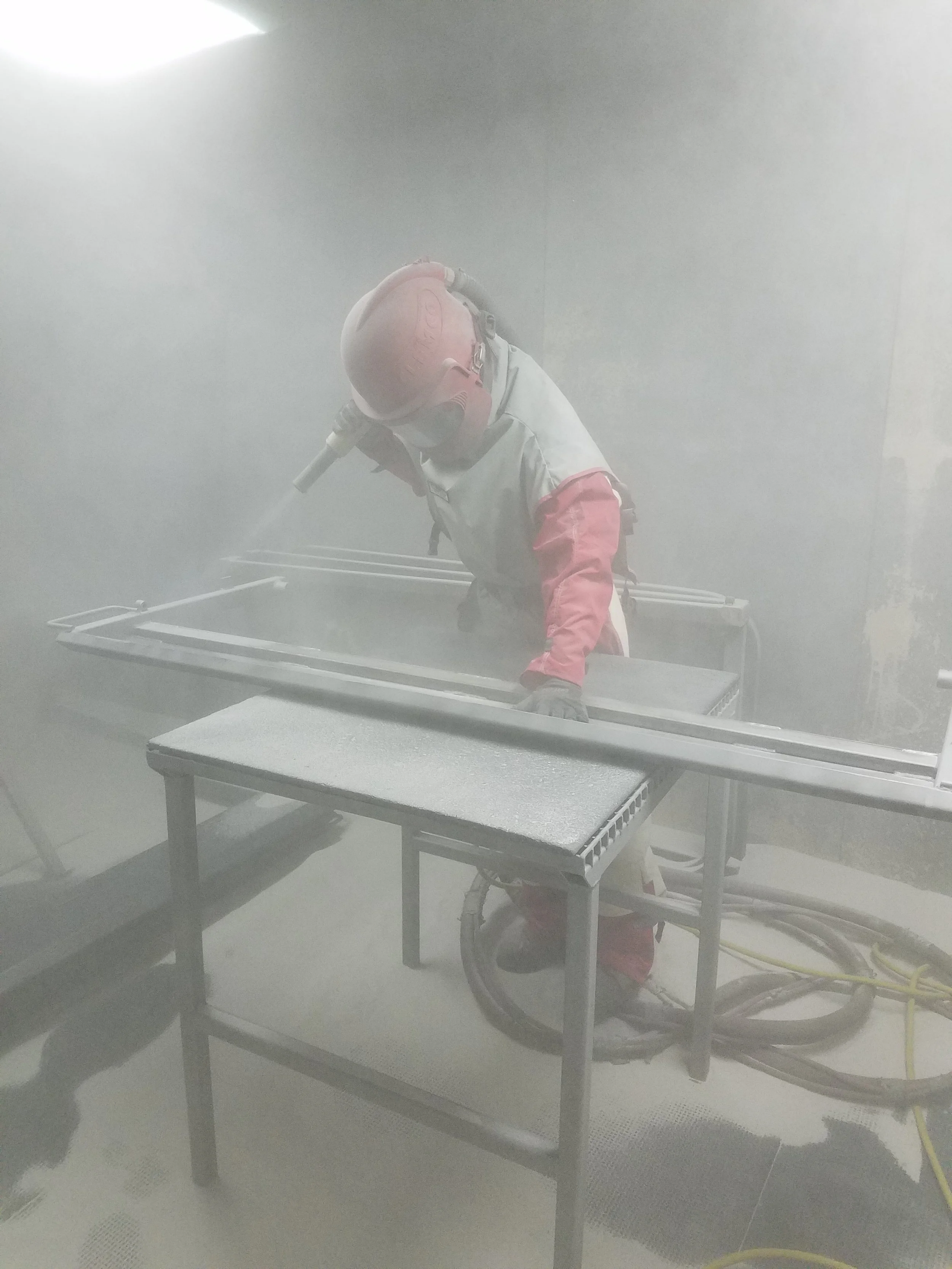 A worker wearing protective gear working in a dusty environment, possibly grinding or sanding a metal frame.
