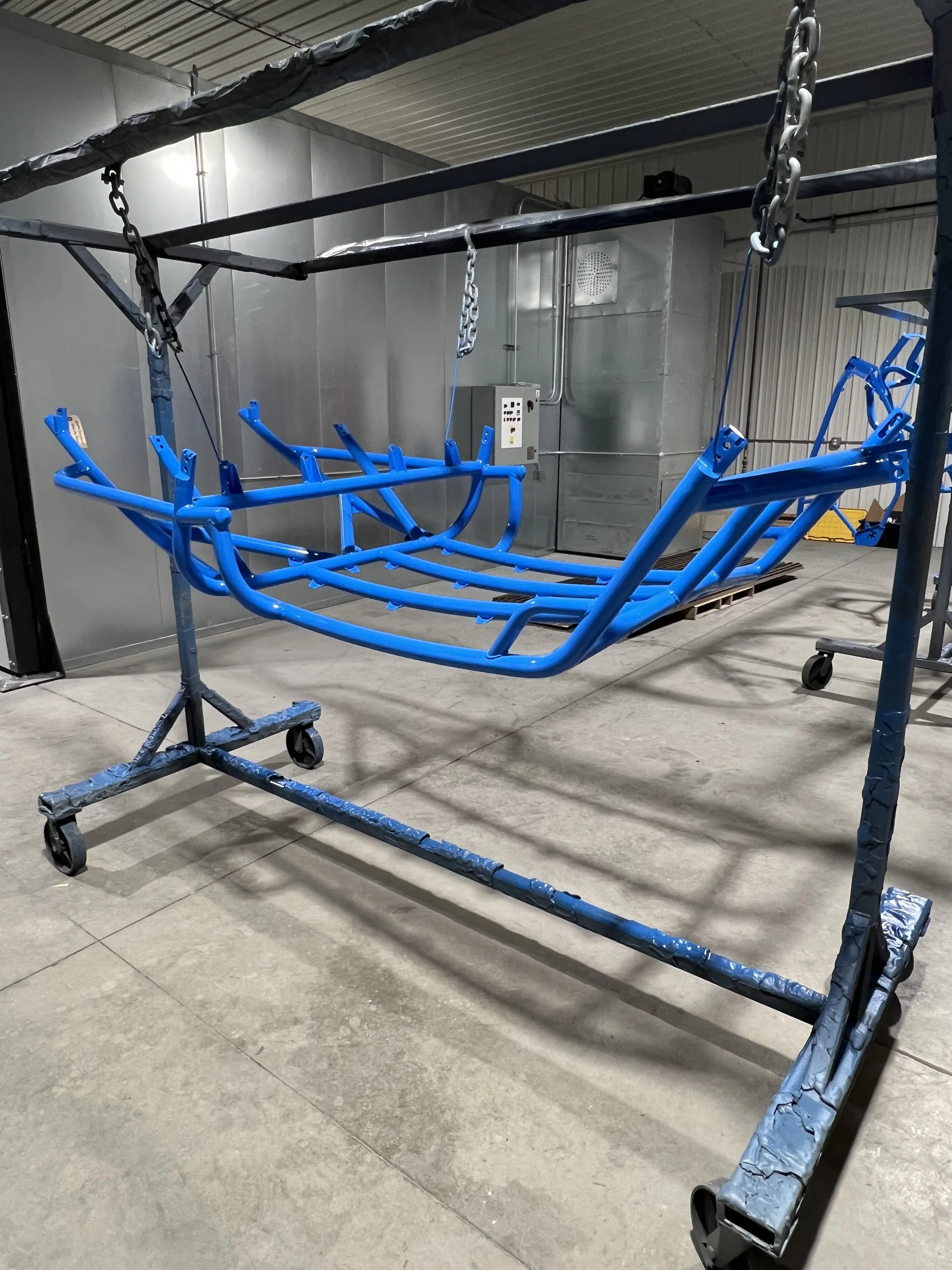 A blue metal frame of a swing suspended by chains inside an industrial or workshop setting, with a gray metal wall and control panel in the background.