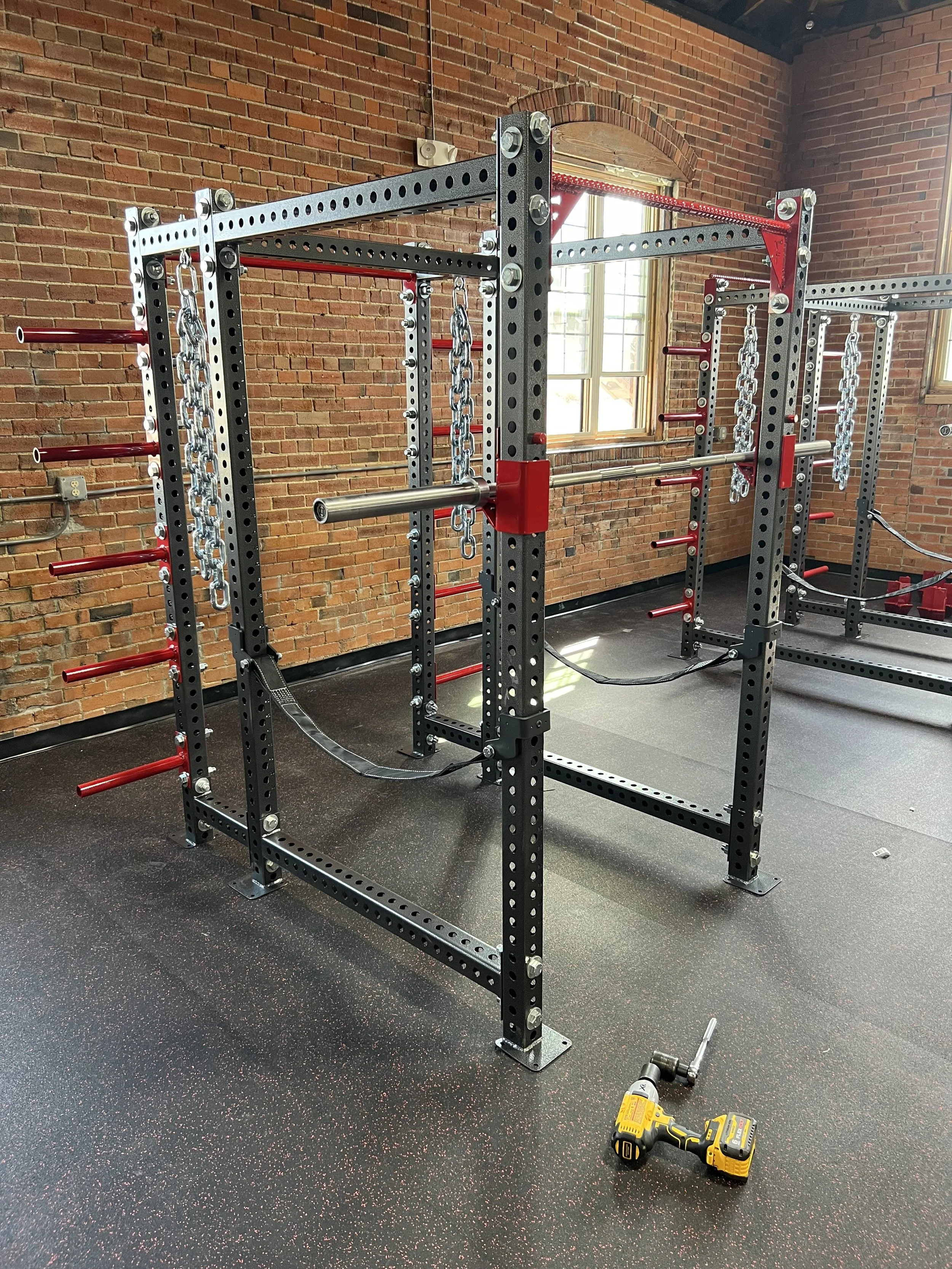 Inside a gym with a metal power rack with red and black accents, chains hanging from the top, and a cordless drill on the floor.