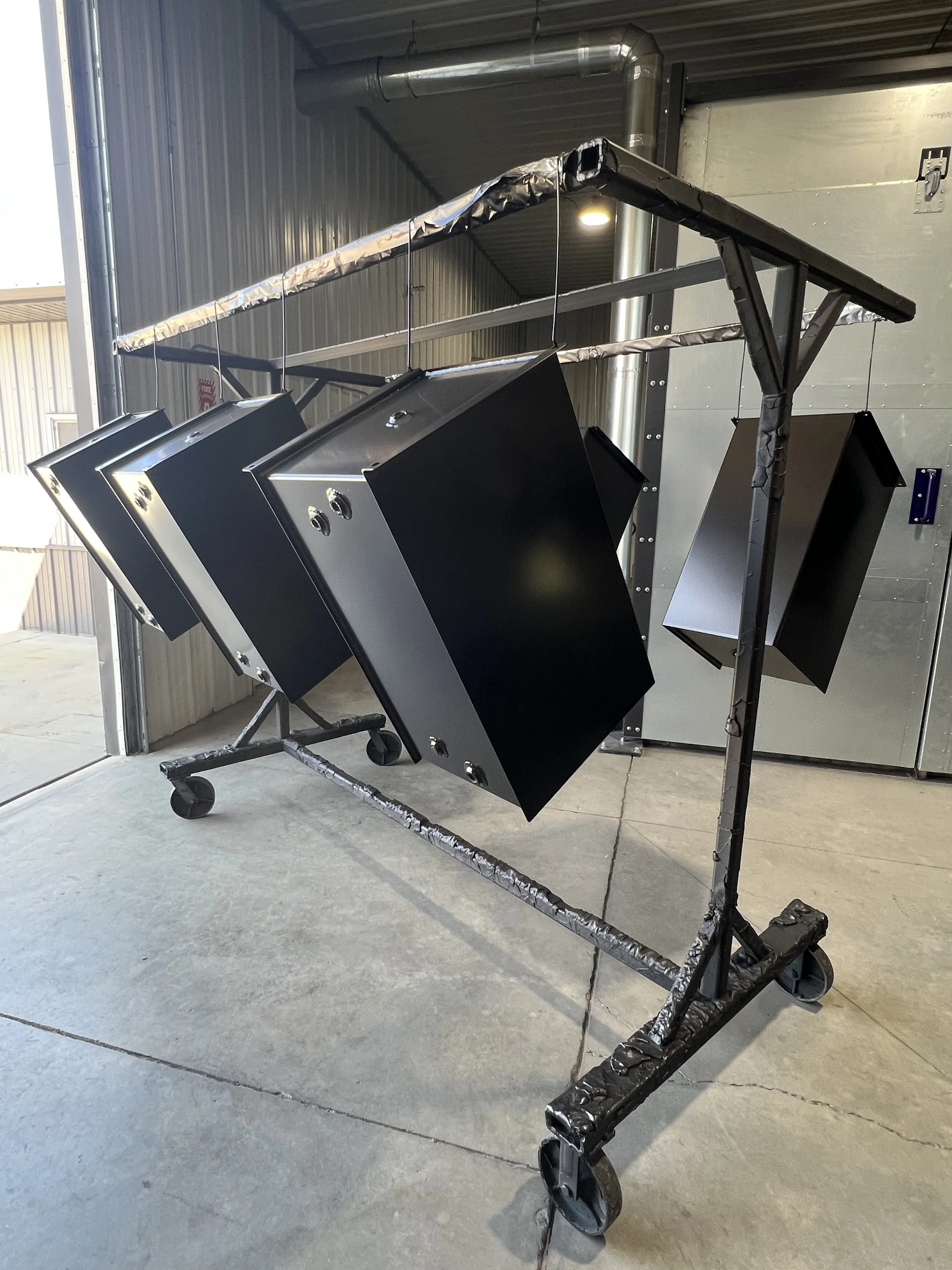 Black metal boxes suspended from a metal frame on caster wheels inside a warehouse or industrial space.