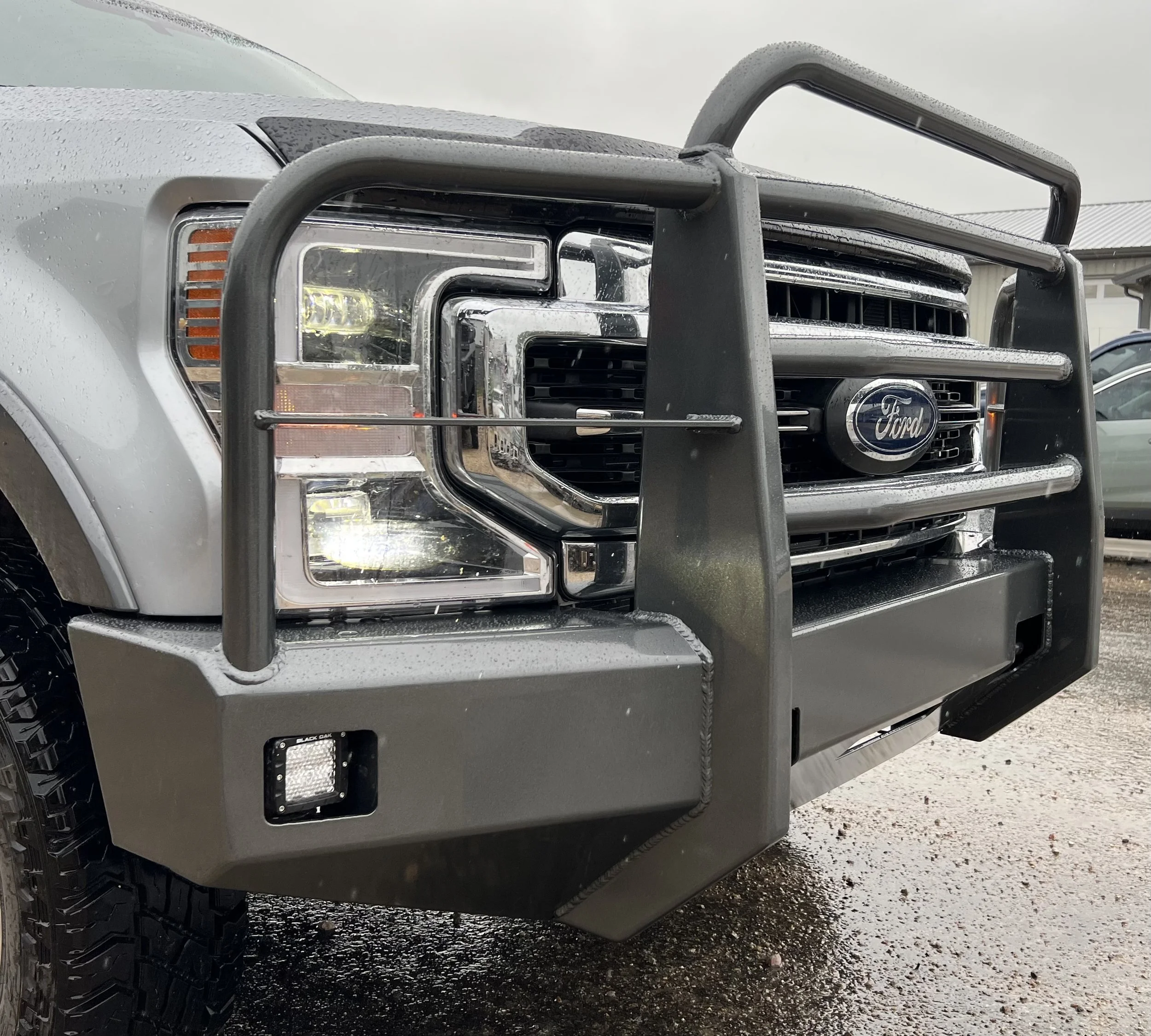 Front view of a silver Ford truck with a black grille guard. The truck has rain droplets on the surface and is parked on a wet ground.