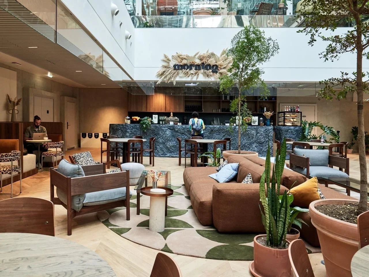 Modern hotel lobby with seating area and a bar in the background. Includes couches, armchairs, indoor plants, and a person working on a laptop.