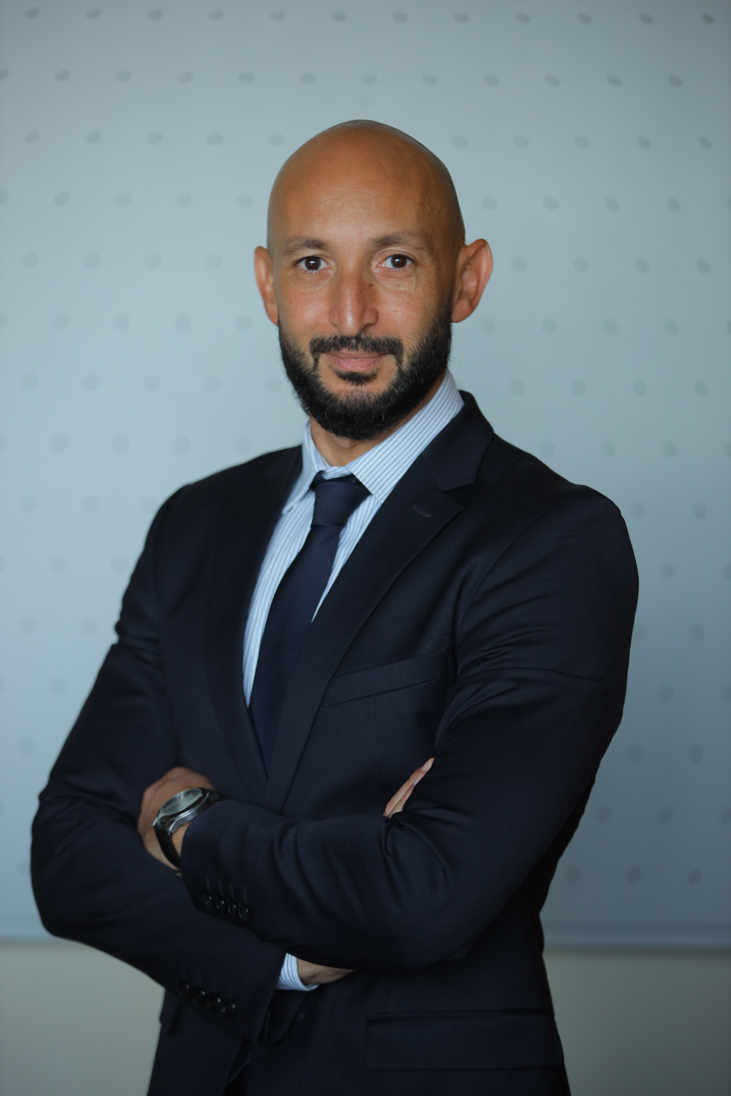 A confident man with a beard in a dark suit and tie, standing with crossed arms in front of a white wall with small gray dots.