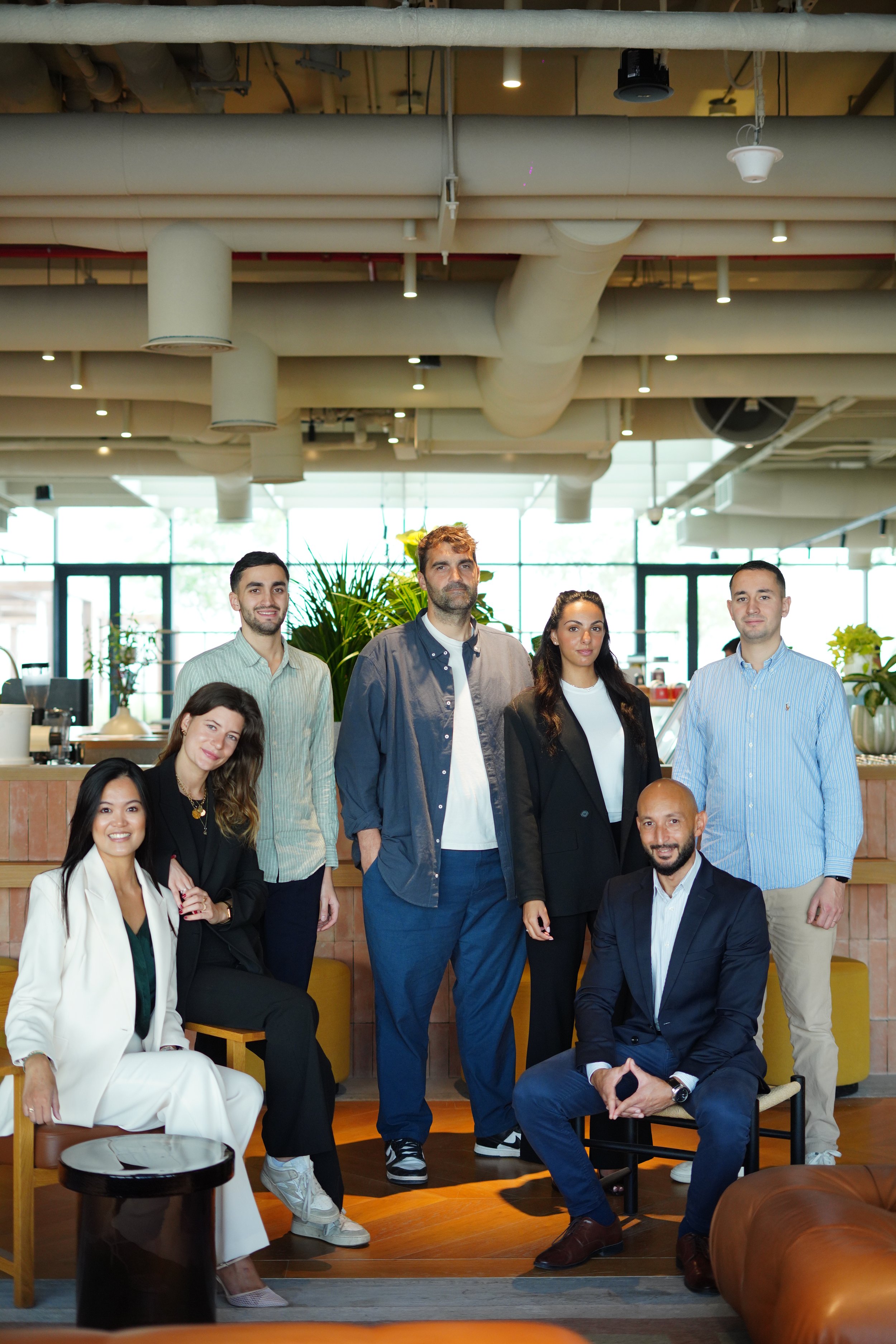 A group of seven diverse professionals posing together in a modern, well-lit office space with large windows, plants, and contemporary furniture.