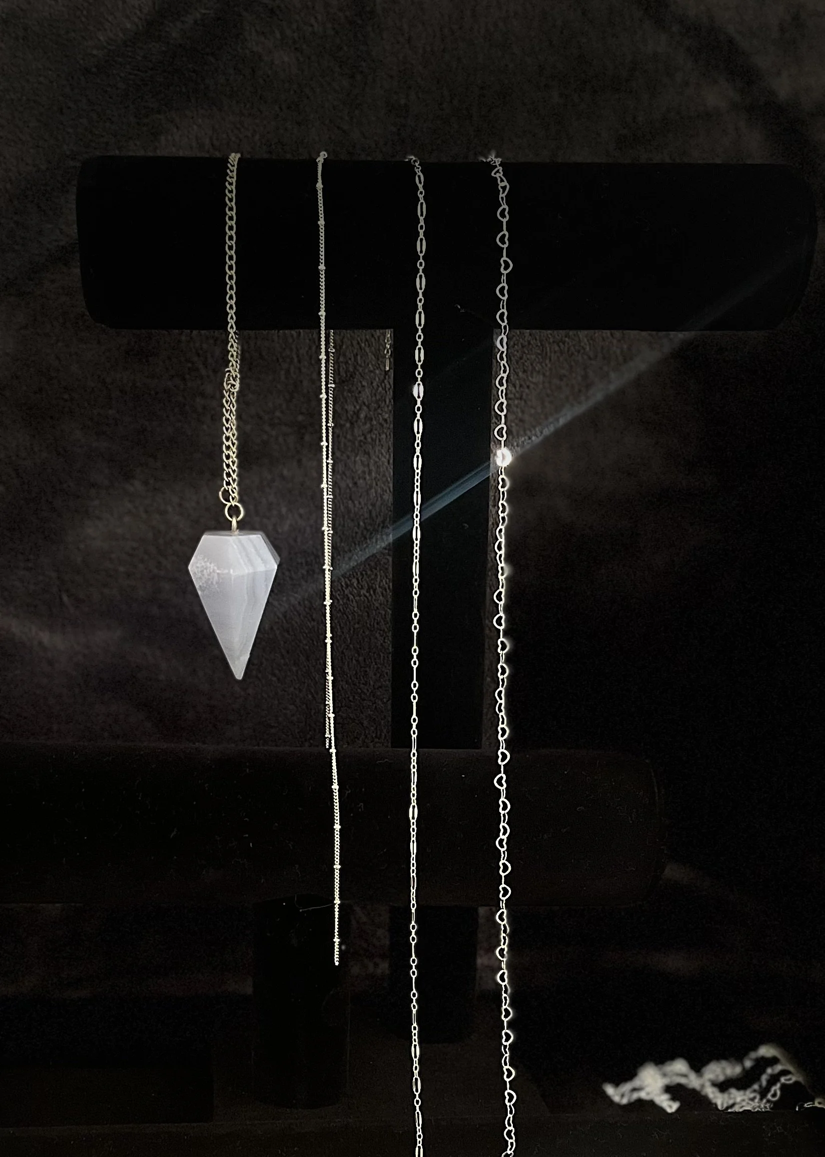 Four silver necklaces hanging on a black display stand, with one featuring a hexagonal white gemstone pendant.