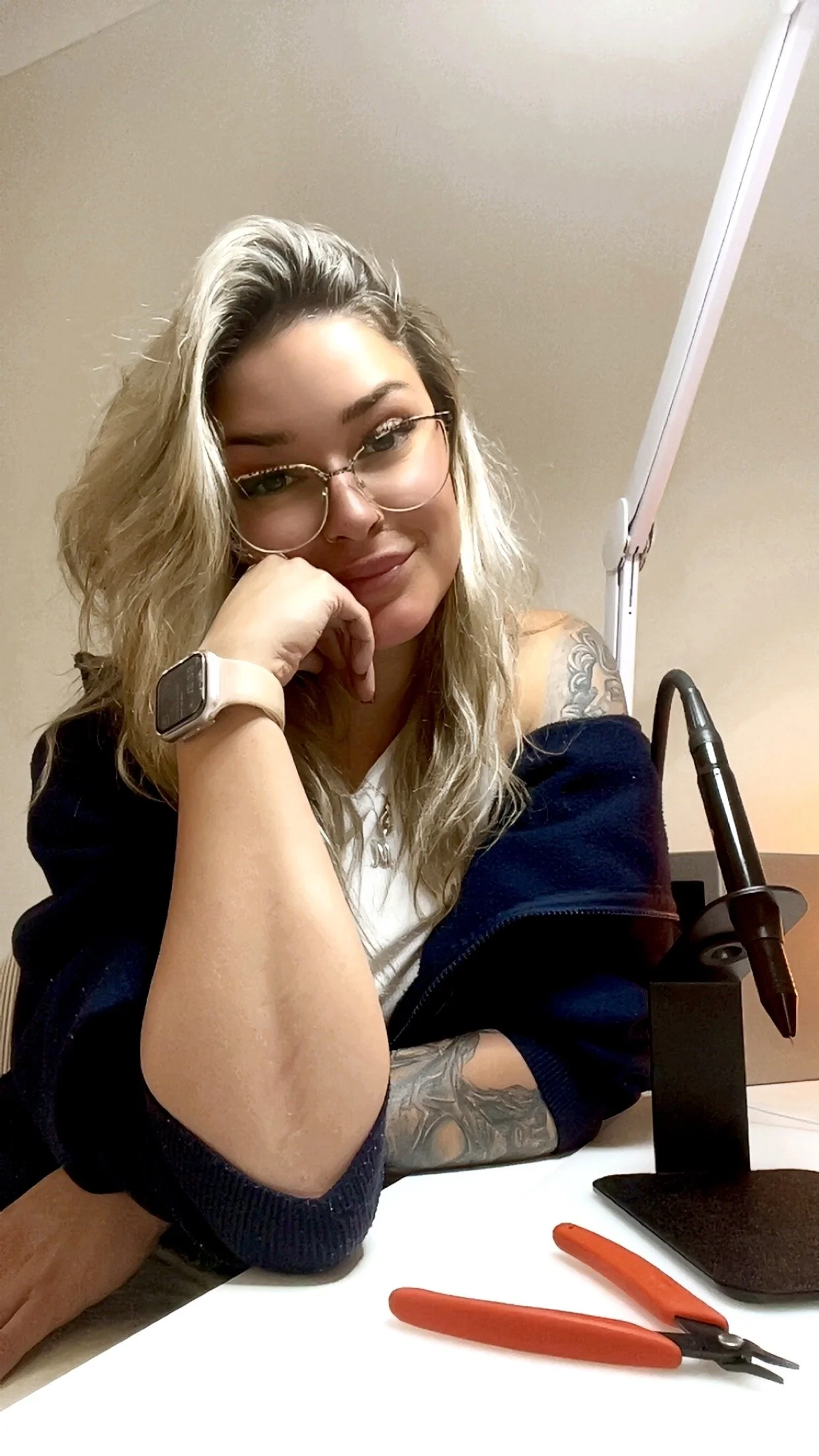 A woman with blonde, wavy hair and glasses is resting her chin on her hand, sitting at a desk with a tattoo on her arm, a smartwatch on her wrist, a soldering iron in a stand, and a pair of orange-handled wire cutters.