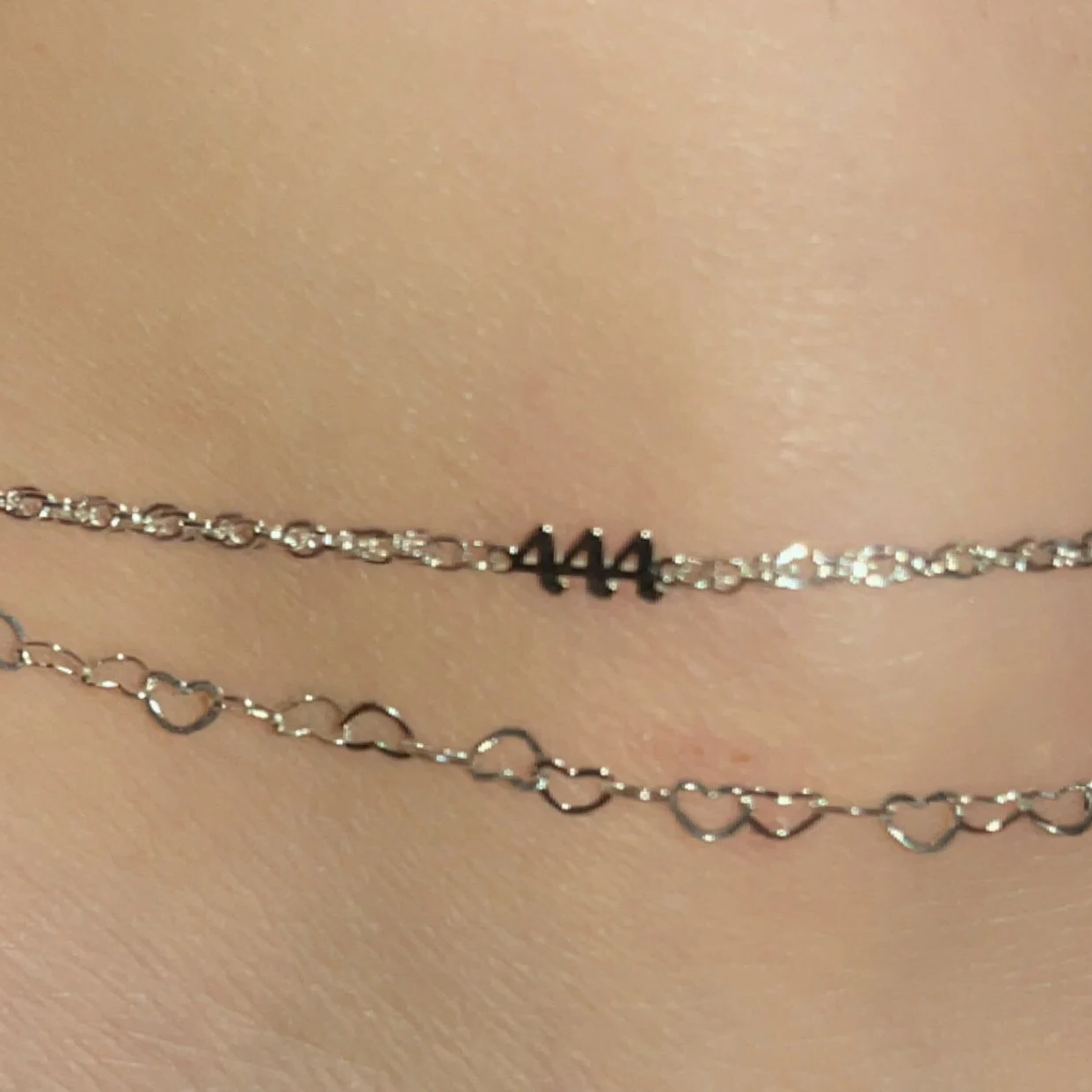 Close-up of two gold necklaces with one featuring the number 444 and the other with small heart-shaped links, worn on smooth skin.