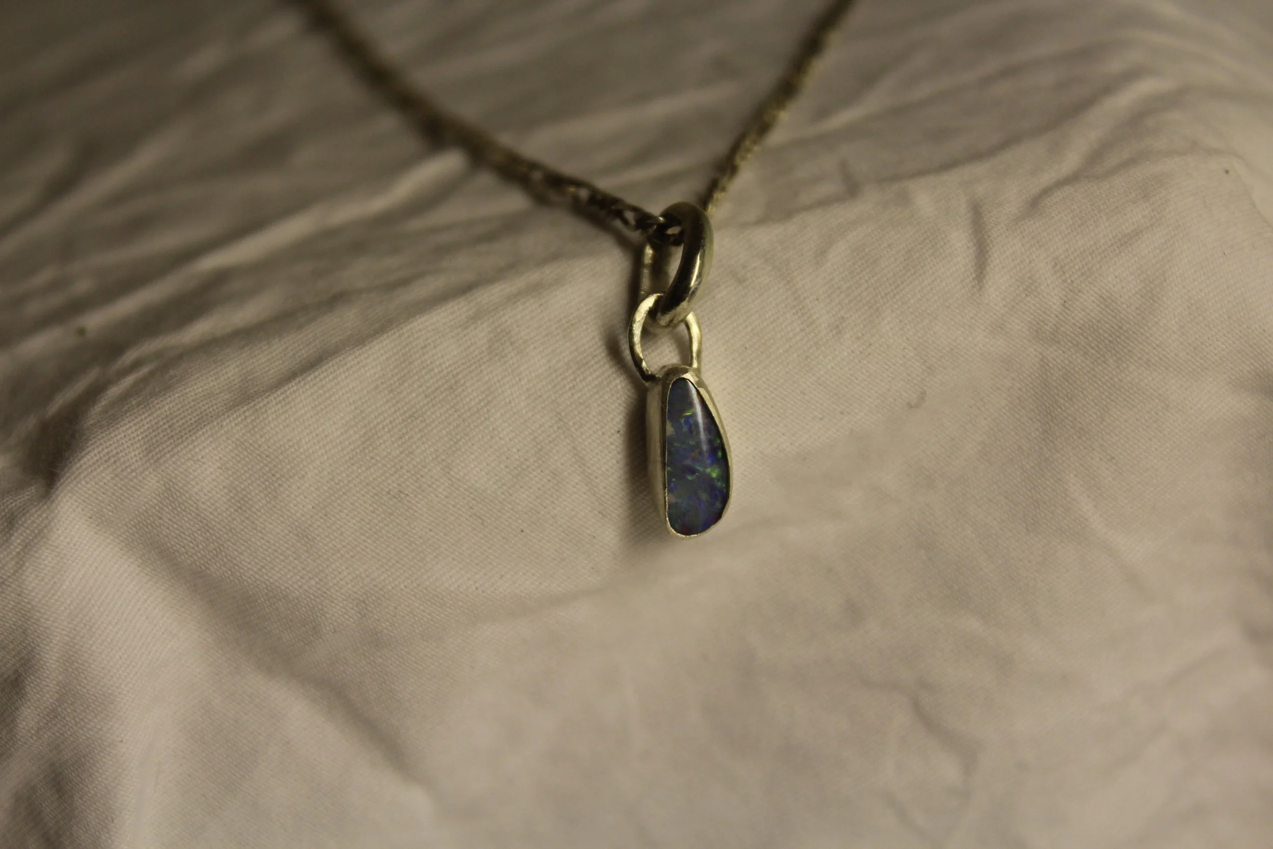 Close-up of a necklace with a pendant, featuring a dark stone with blue and green highlights, resting on a white fabric surface.