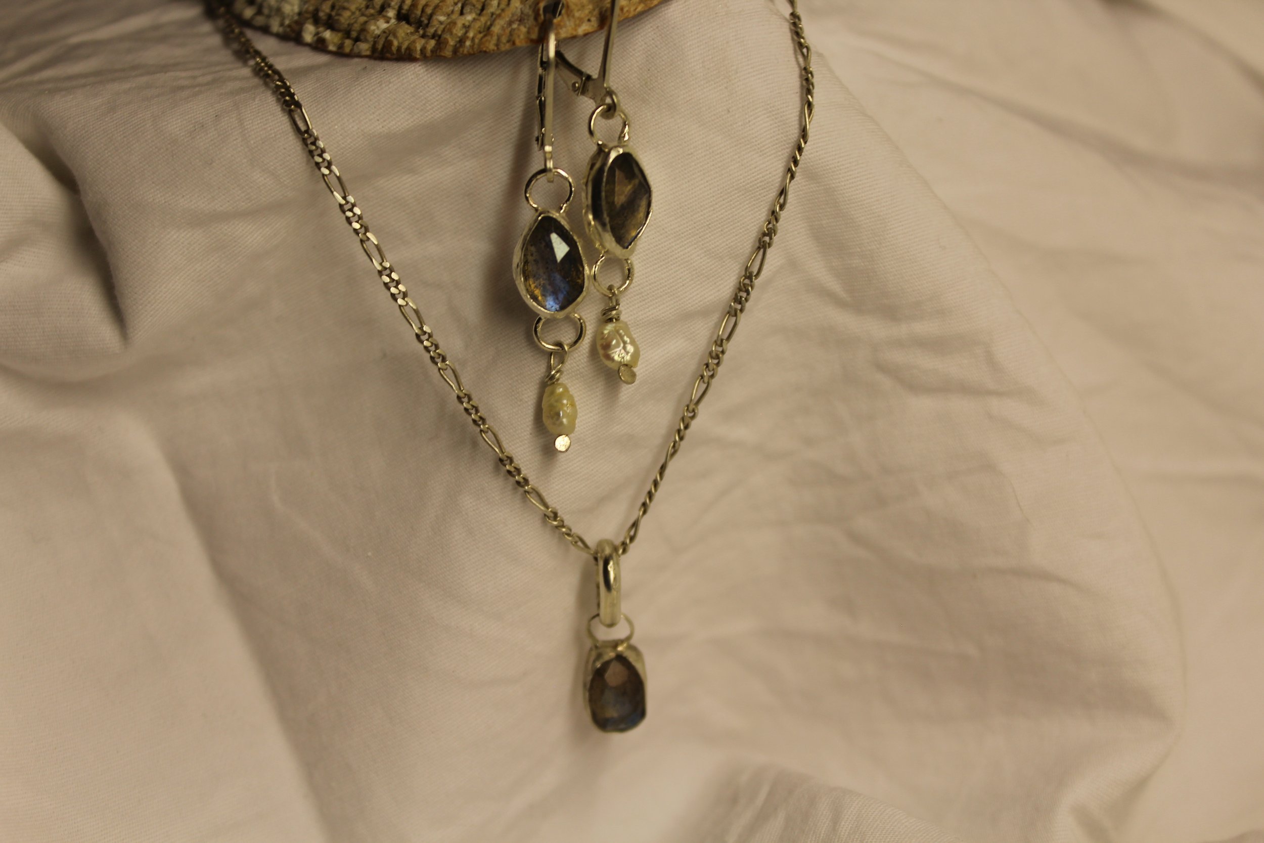 Silver jewelry including layered chains and pendants with dark gemstones and pearls, displayed against light fabric.