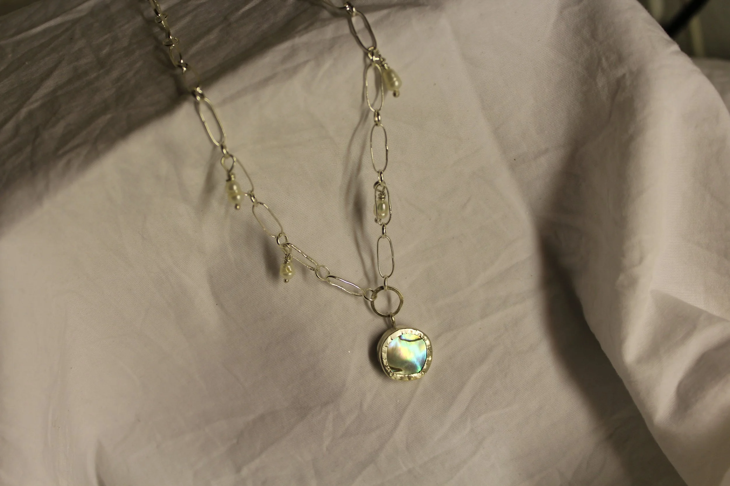 A silver chain necklace with small pearl accents and a square pendant containing a colorful, iridescent shell piece, photographed against light-colored fabric.