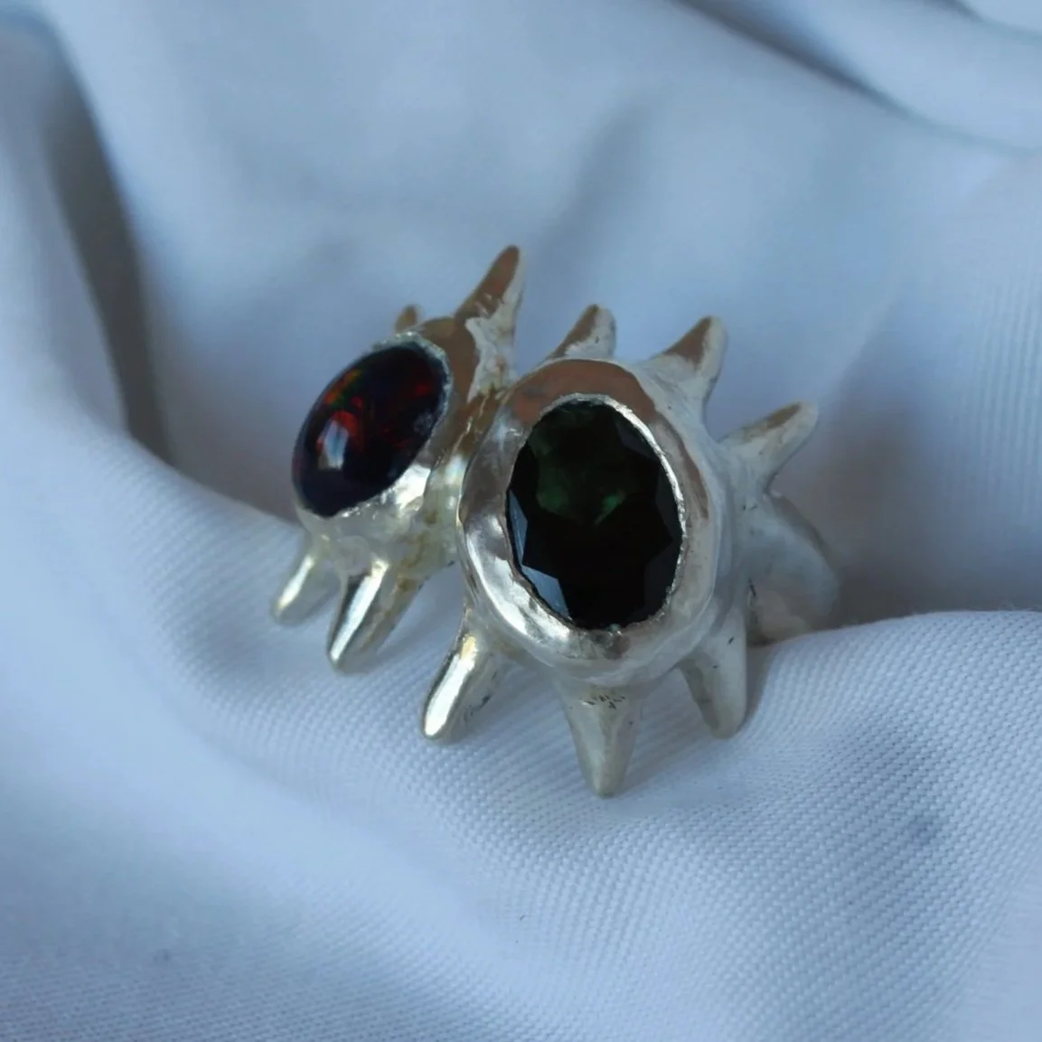 A pair of silver rings with black gemstones, designed with spikes and black accents, resting on a white fabric background.