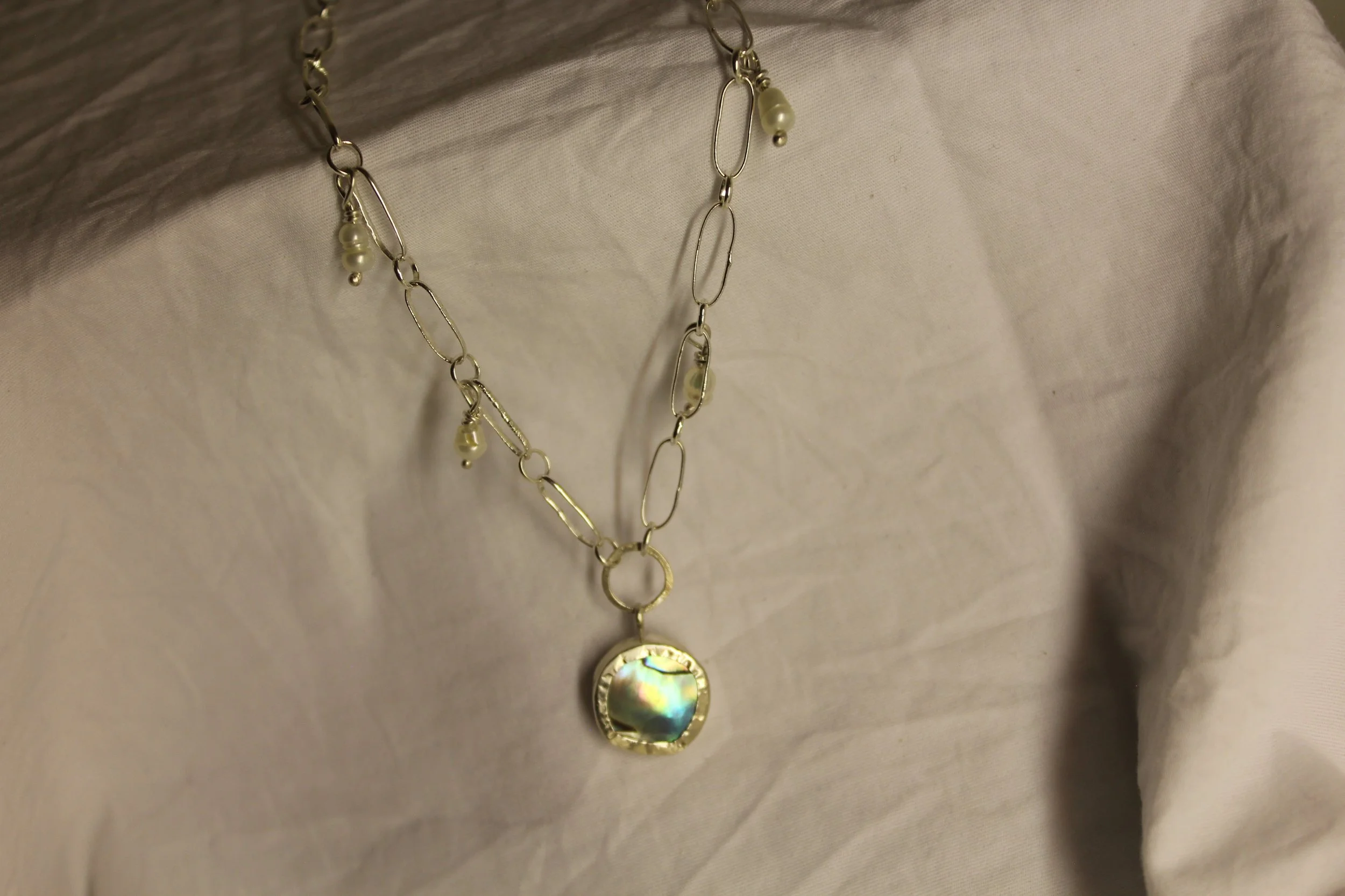 A silver necklace with links and small pearl charms, featuring a pendant with a round, iridescent mother-of-pearl setting.