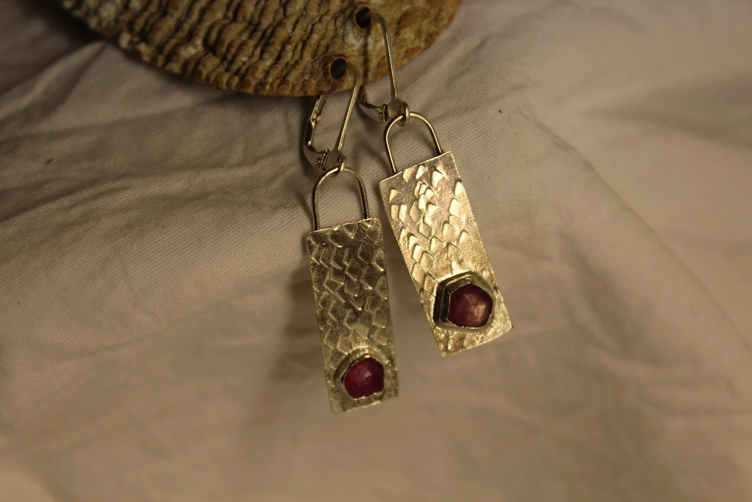 Gold textured rectangular earrings with red stones at the bottom hanging from metallic hooks, resting on fabric near a piece of wood.