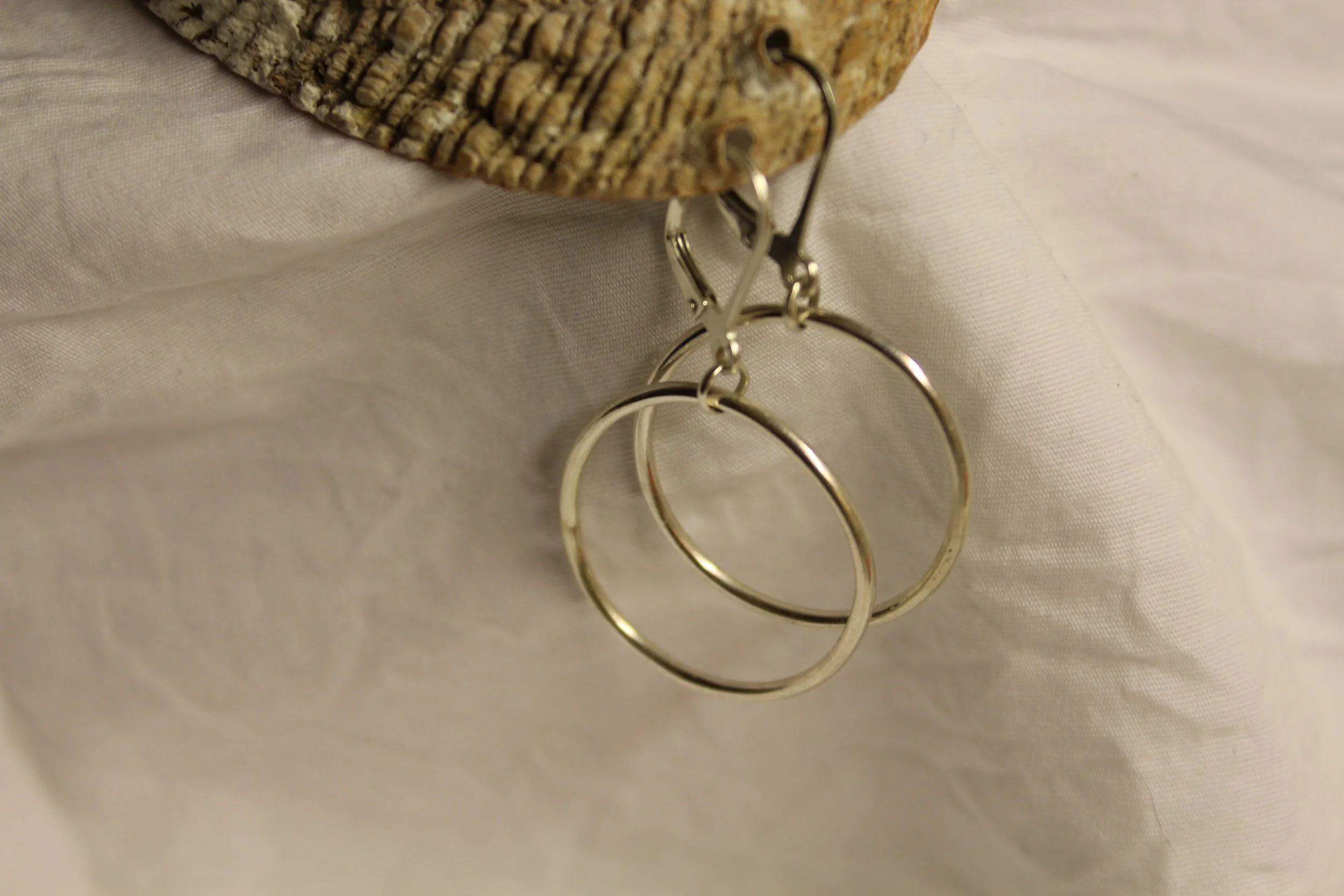 Silver hoop earrings hanging from a brown textured piece of wood or shell, against a light fabric background.