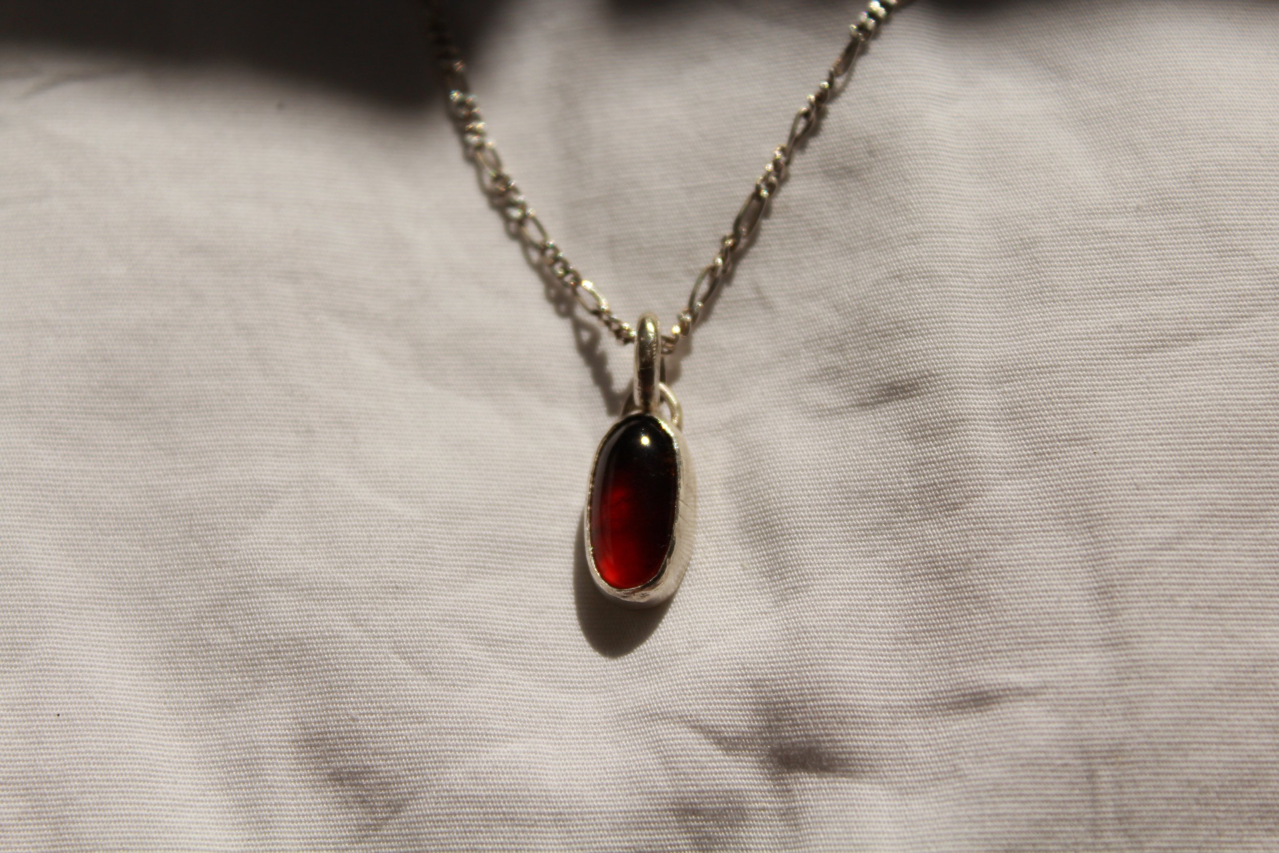 A silver chain necklace with a red oval pendant, resting on a white fabric background.