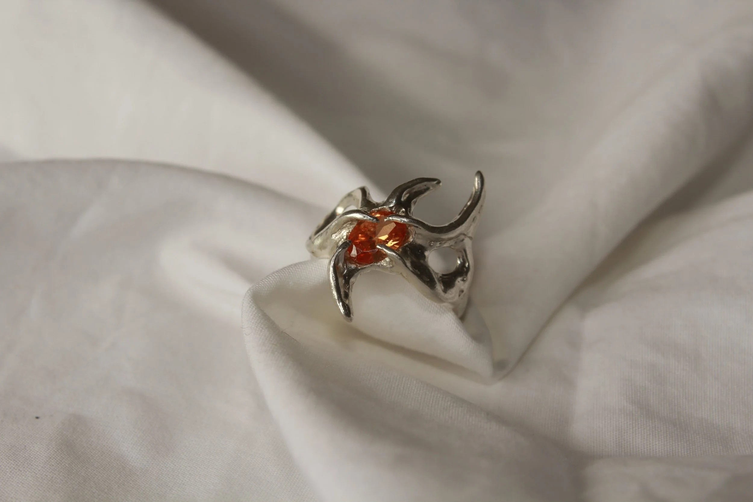 A silver ring with a design resembling a bat, featuring orange gemstones in the center, resting on white fabric.