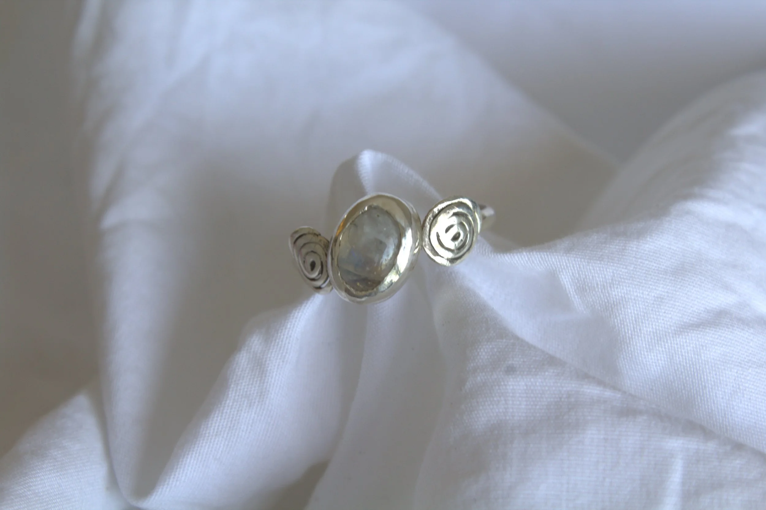 Silver ring with a central clear gemstone, featuring spiral designs on either side, resting on white fabric.