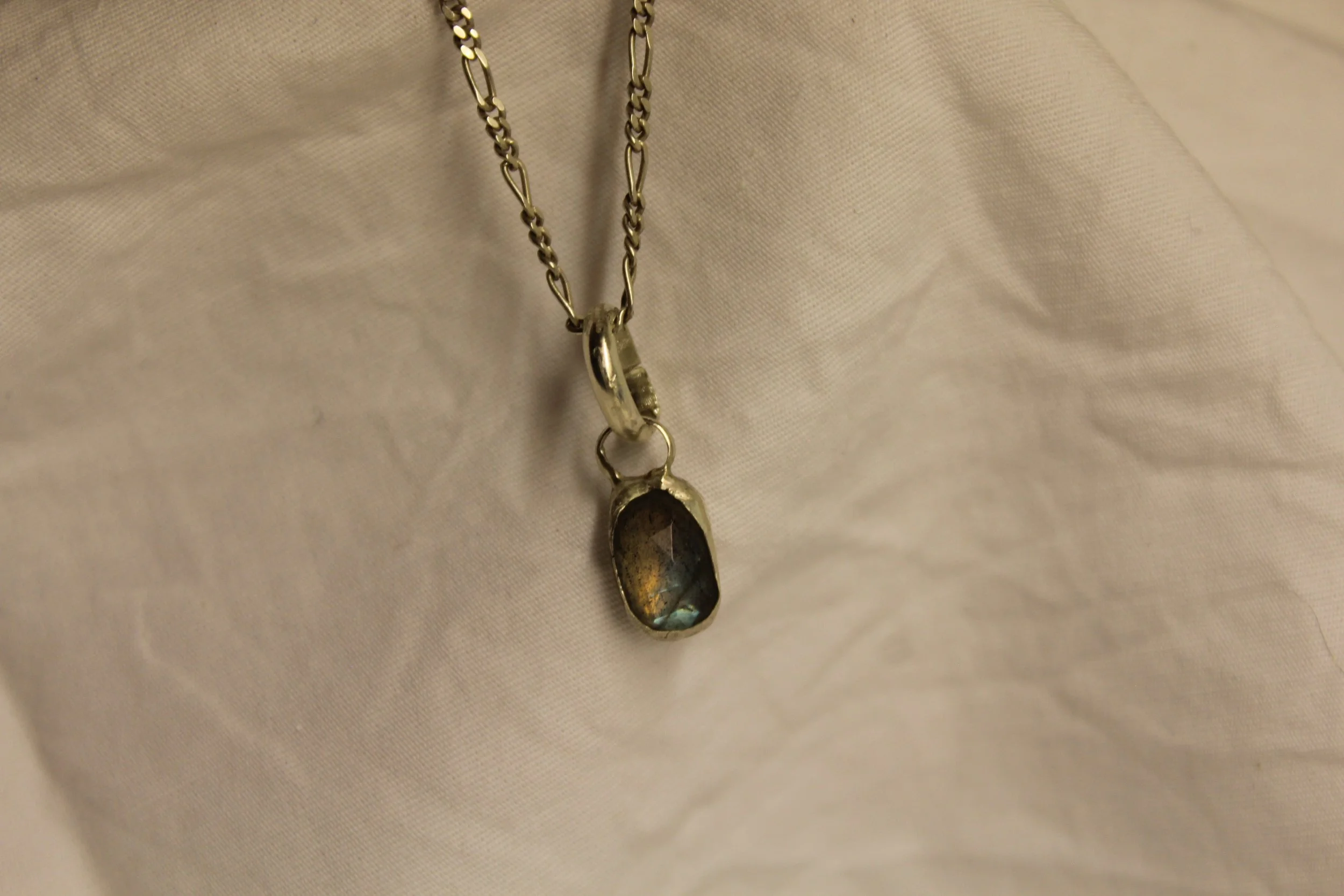 A silver necklace with a rectangular, iridescent gemstone pendant, resting on beige fabric.