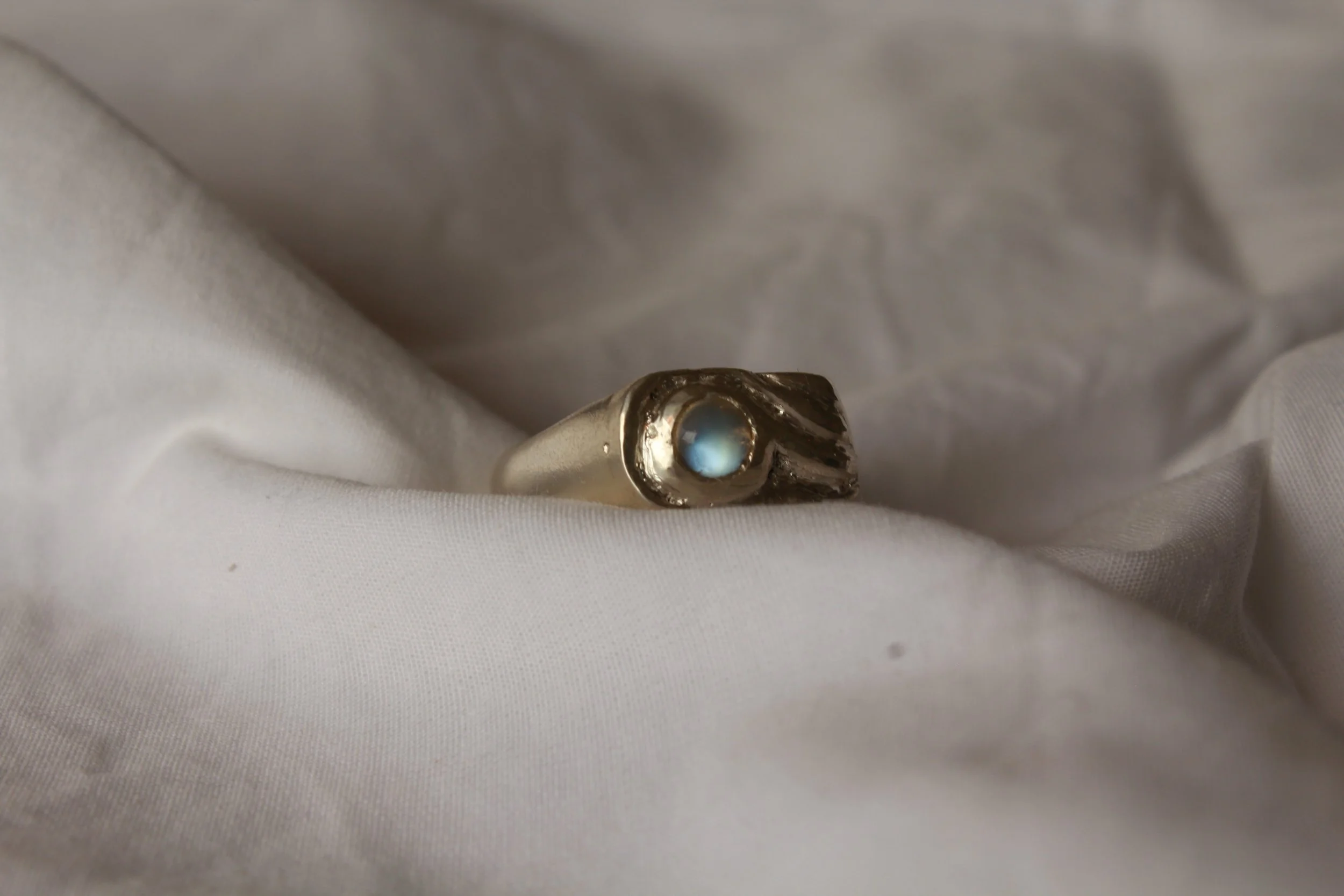 A silver ring with a round, blue gemstone set in a textured, irregular band, resting on white fabric.