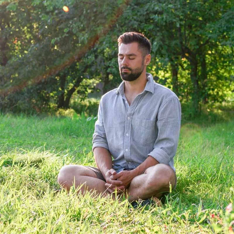 A man sitting cross-legged on the grass with eyes closed, surrounded by trees and greenery, in a peaceful outdoor setting.