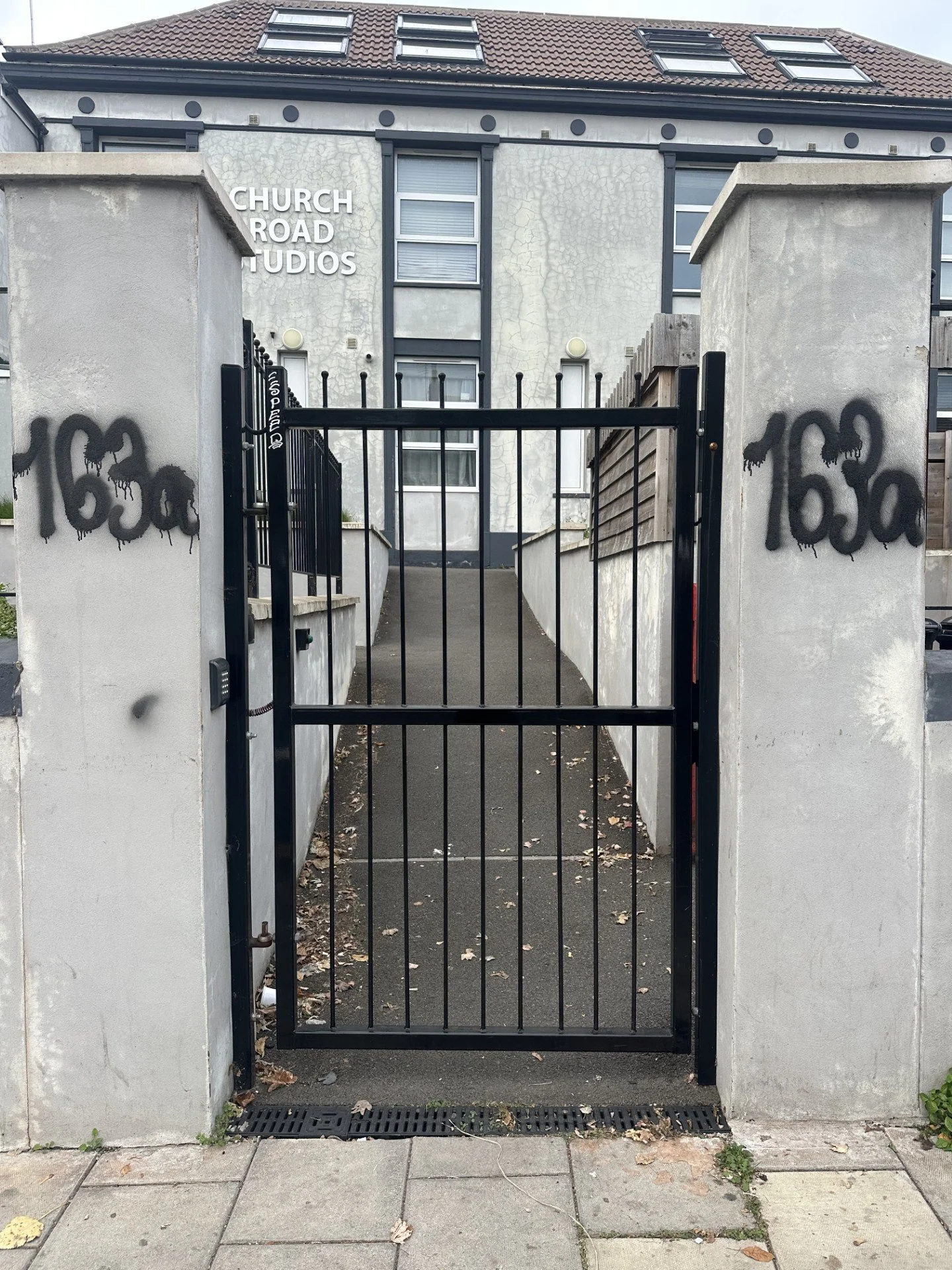A black metal gate with a latch in front of a driveway leading to a building with signs that read 'Church Road Studios.' The building has a white facade with visible cracks, gray window frames, and a brown tiled roof with skylights. There are black graffiti tags on the concrete pillars on either side of the gate.