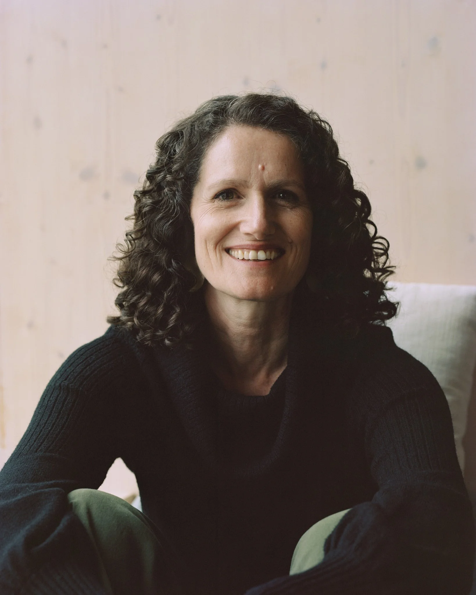 A woman with curly hair wearing a sweater, sitting outdoors with wooden stairs in the background.