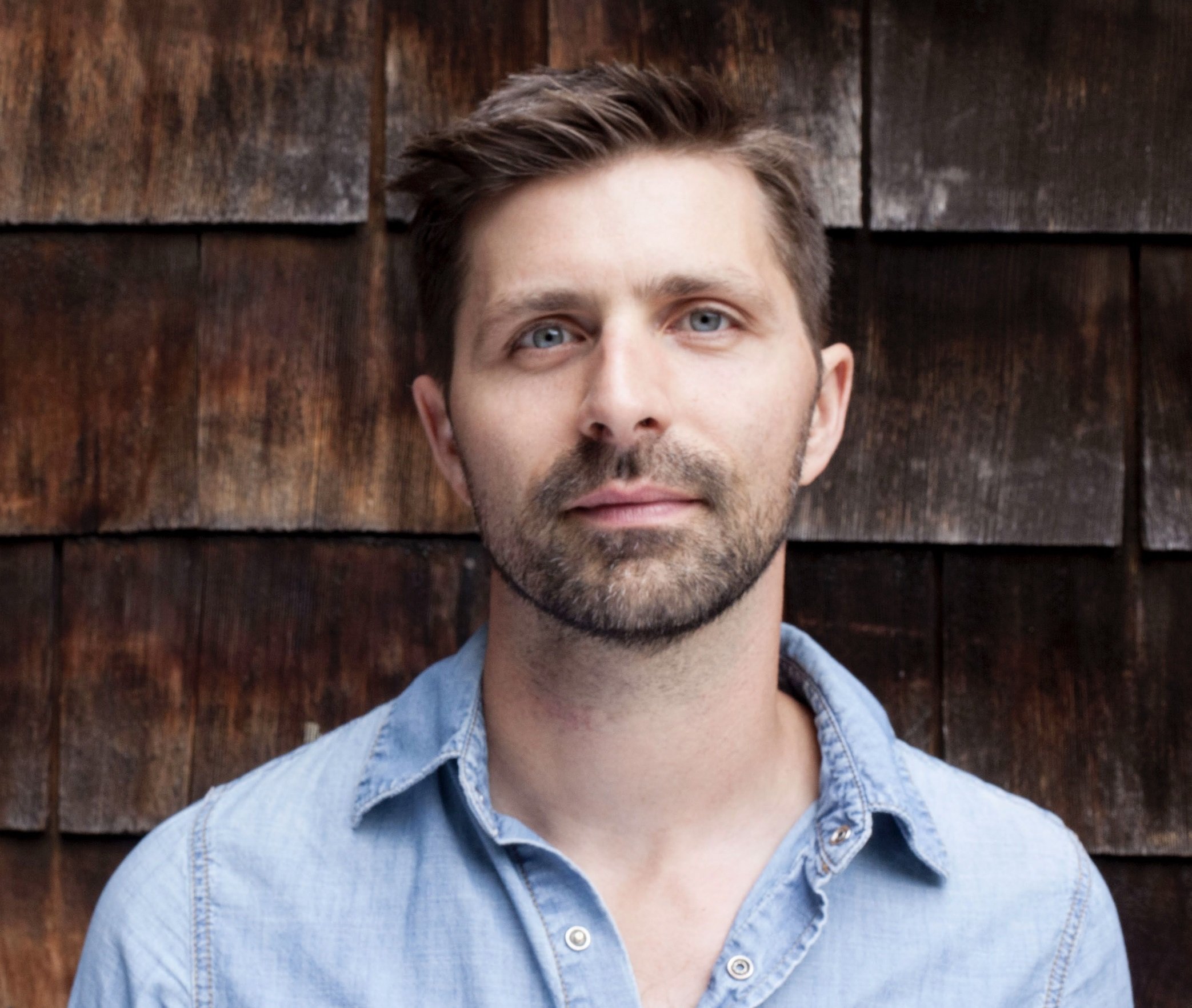 A portrait of a man with short brown hair and blue eyes, wearing a light denim shirt, standing outdoors against a rustic wooden background.