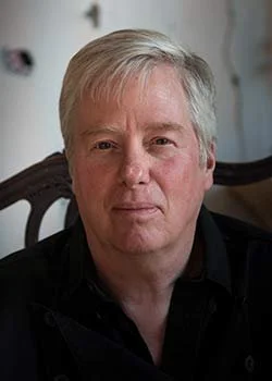 A middle-aged man with gray hair and fair skin, sitting indoors with a blurred background.