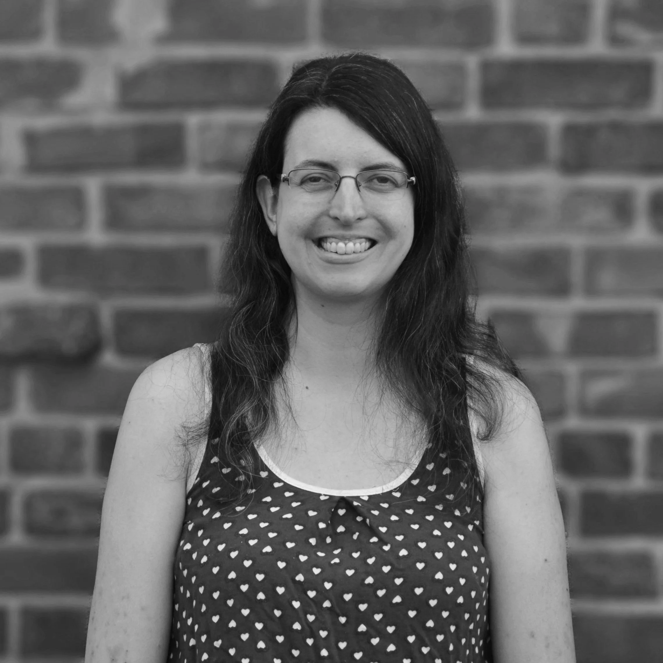A woman with glasses and long dark hair smiling in front of a brick wall, wearing a sleeveless top with a heart pattern.