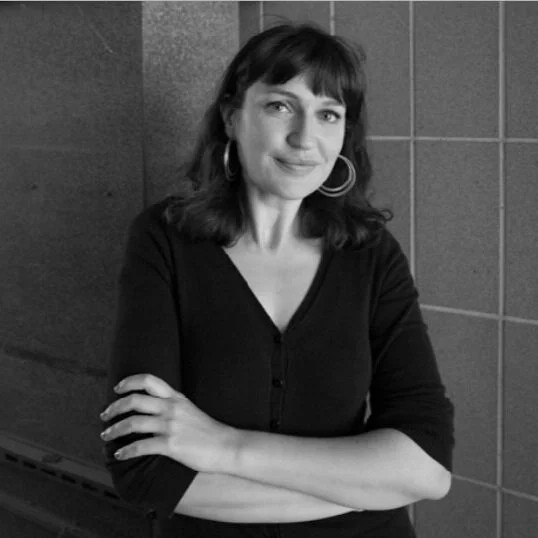 Black and white photo of a woman with shoulder-length dark hair, wearing hoop earrings and a black top, standing against a tiled wall.